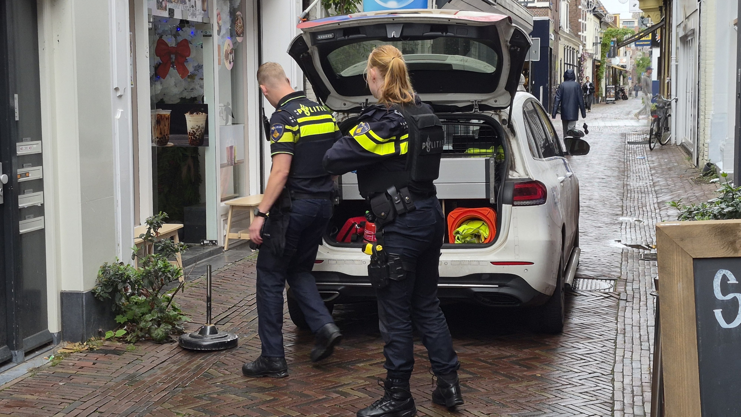 Twee politieagenten staan bij een politieauto met open achterklep in een smalle straat bij een winkelpand