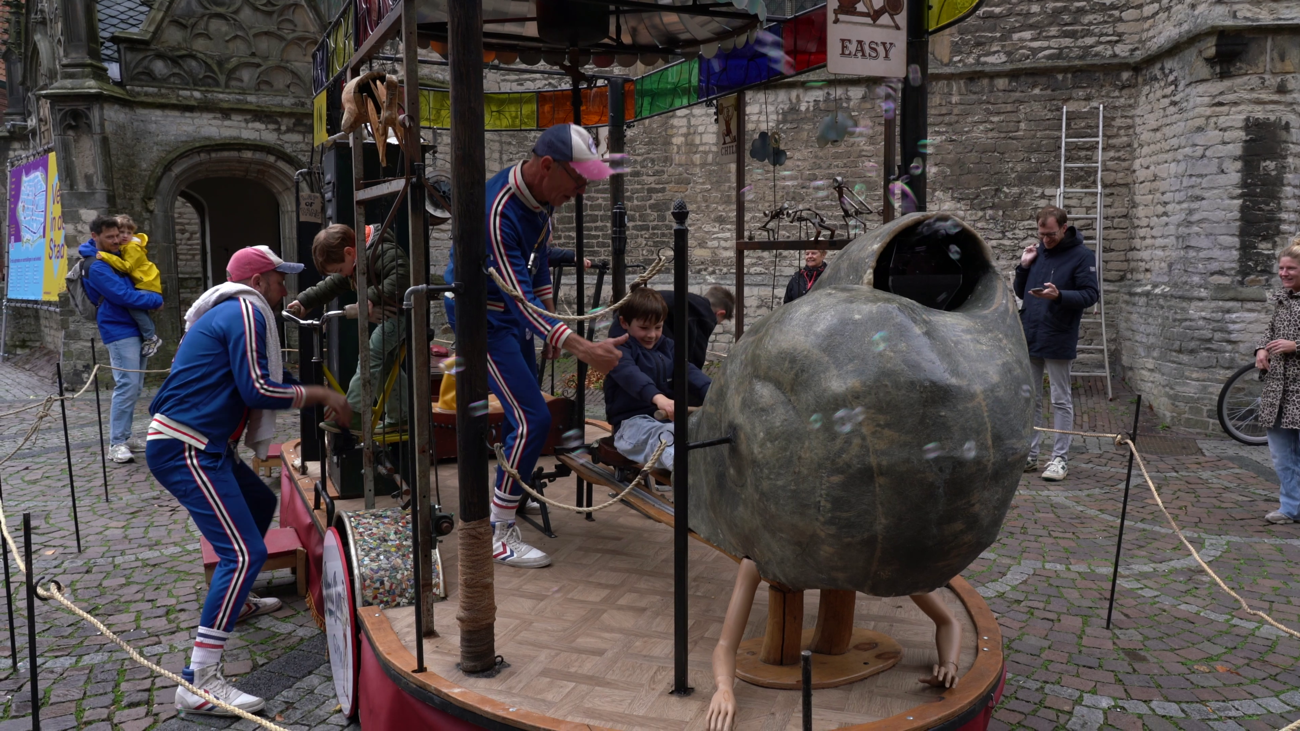 Straatact met twee performers in blauwe pakken die kinderen begeleiden bij een draaiende kermisattractie met grote bol