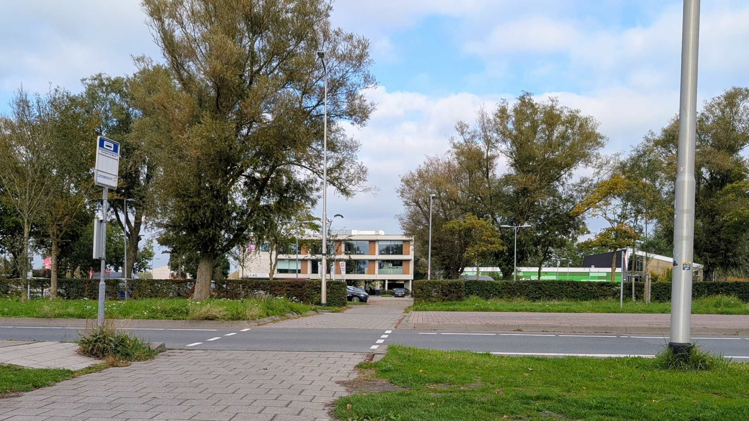 Bushalte bij een rustige straat met trottoir, bomen en een kantoorachtig gebouw op de achtergrond onder een deels bewolkte lucht