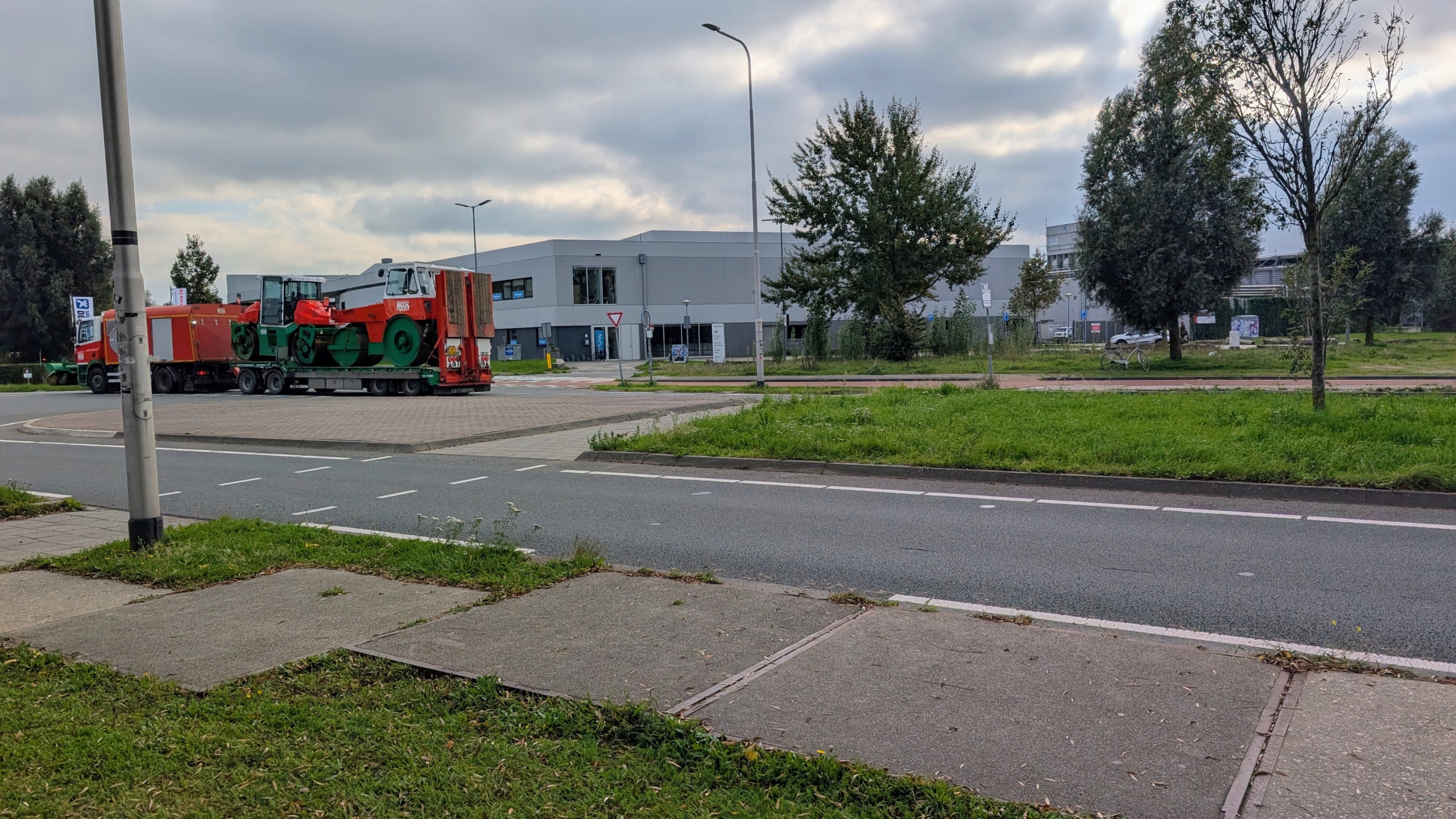 Vrachtwagen met oplegger vervoert een grote wegwals langs een rotonde bij grijze bedrijfsgebouwen