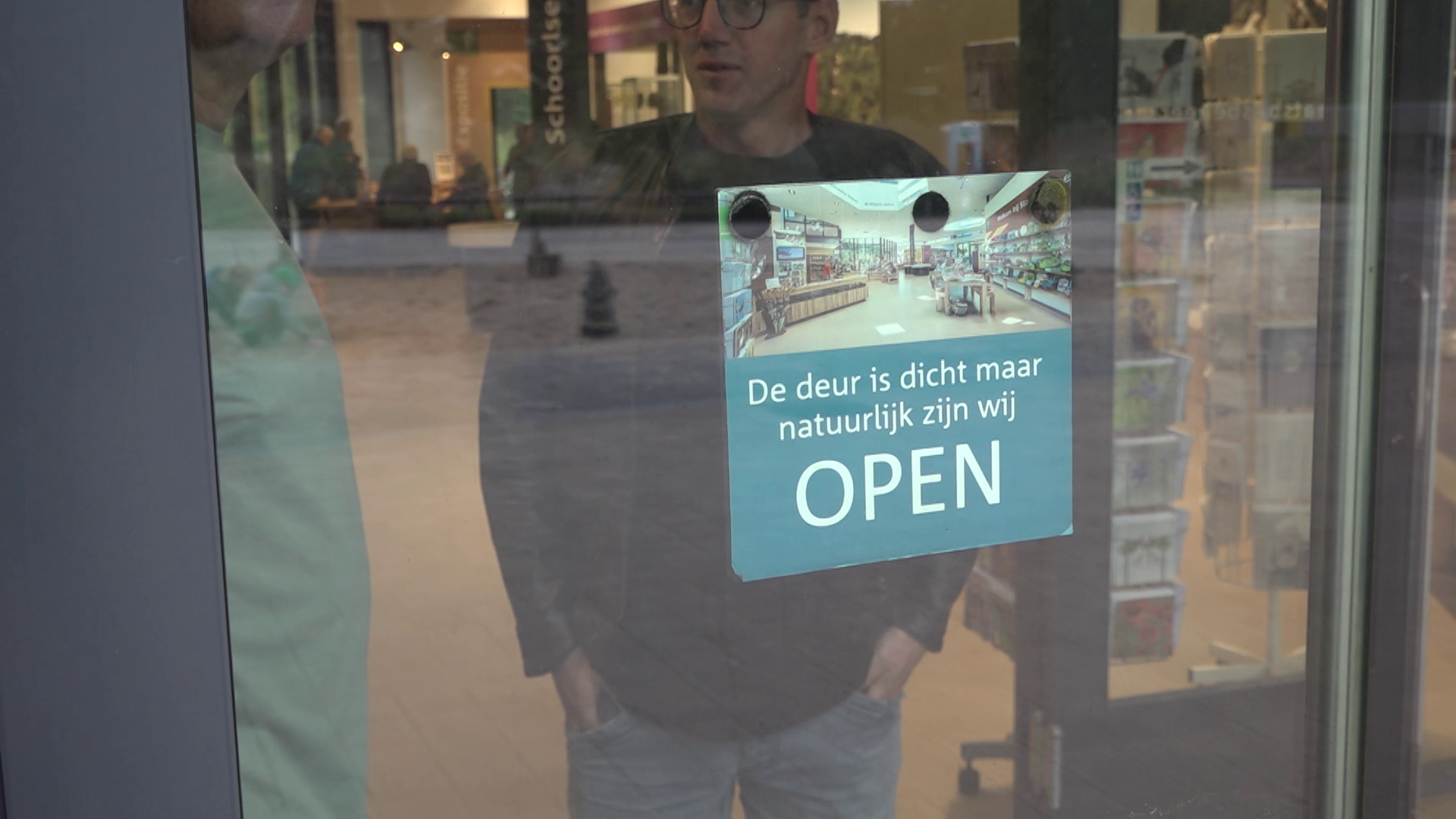 Briefje op glazen deur met tekst De deur is dicht maar natuurlijk zijn wij OPEN, met reflectie van twee personen