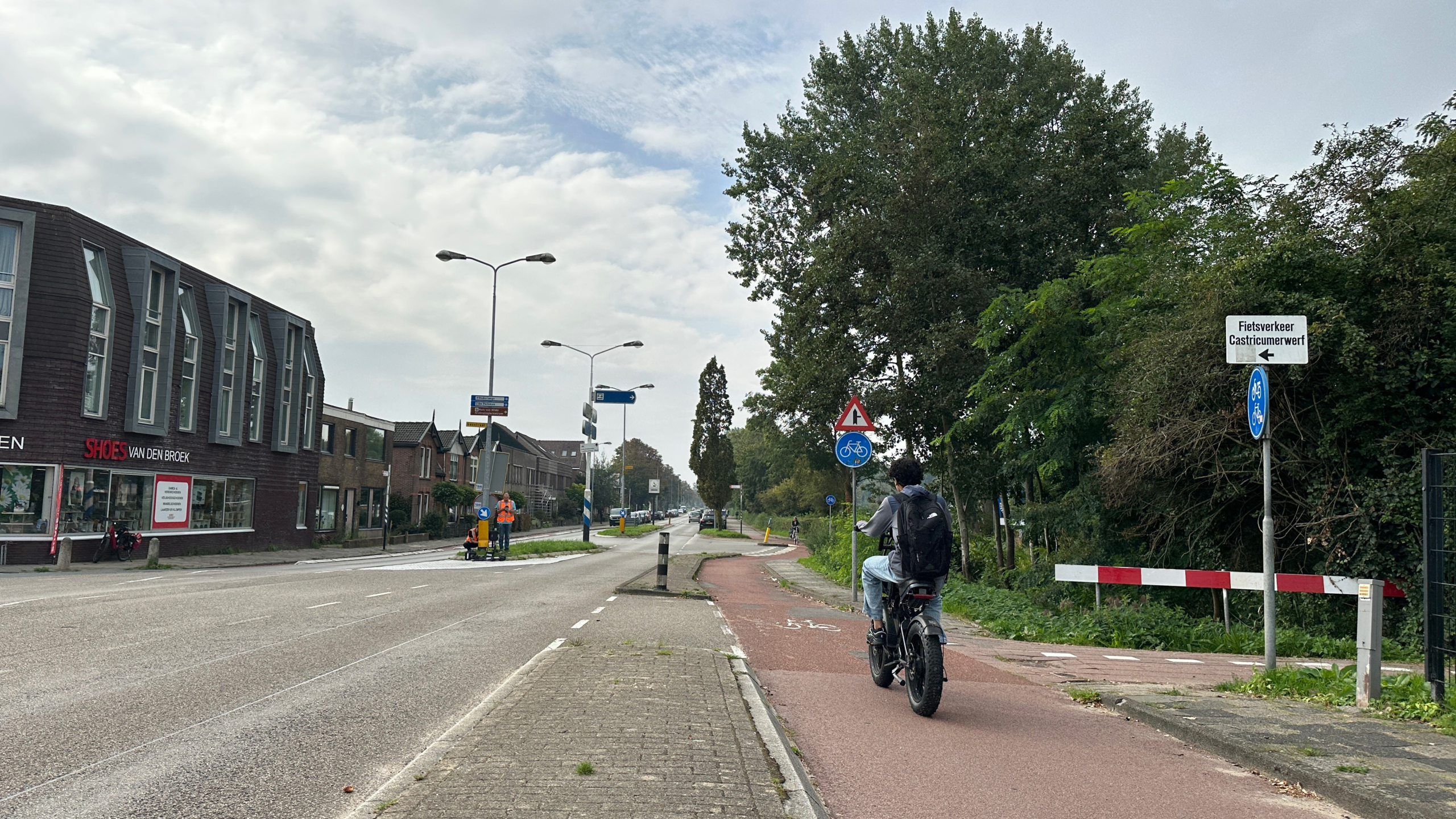 Fietser met rugzak op rood fietspad langs een weg met winkels en bomen, bij een bord richting Castricummerwerf