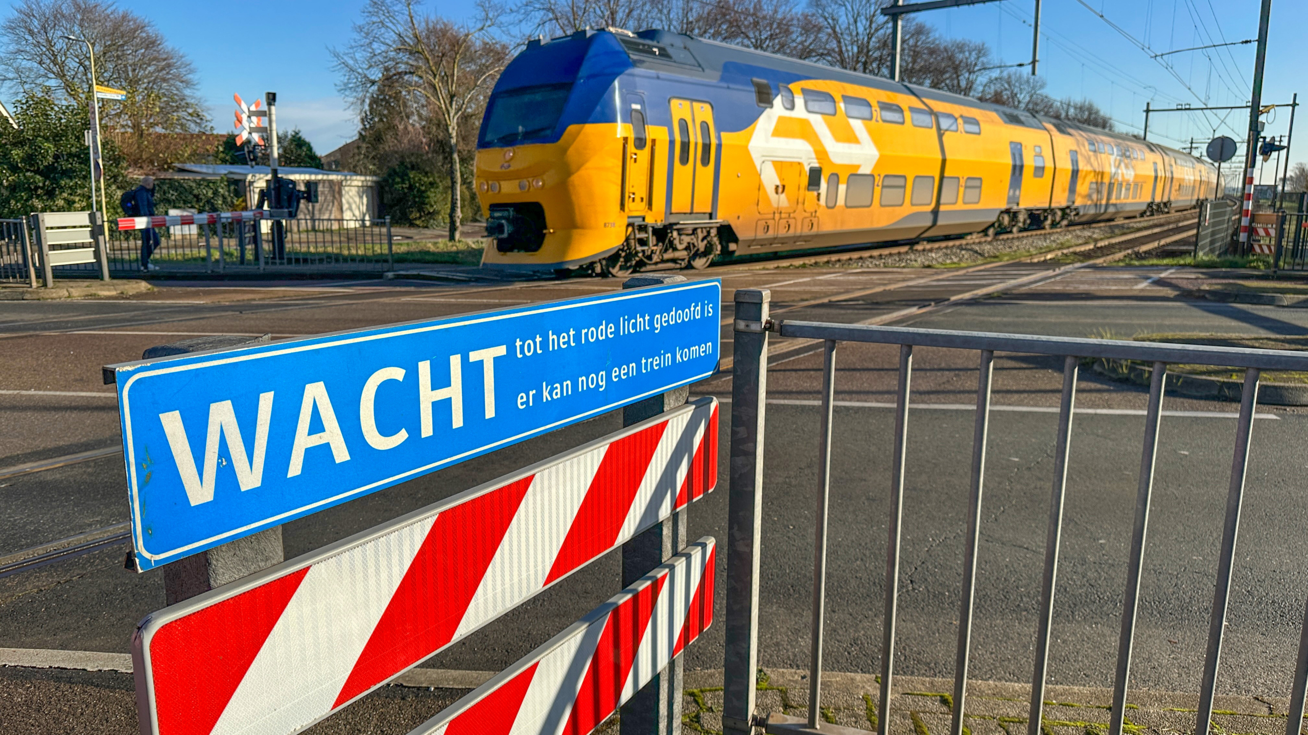 Gele dubbeldekker trein rijdt langs een spoorwegovergang met hekwerk en bord WACHT, tot het rode licht gedoofd is