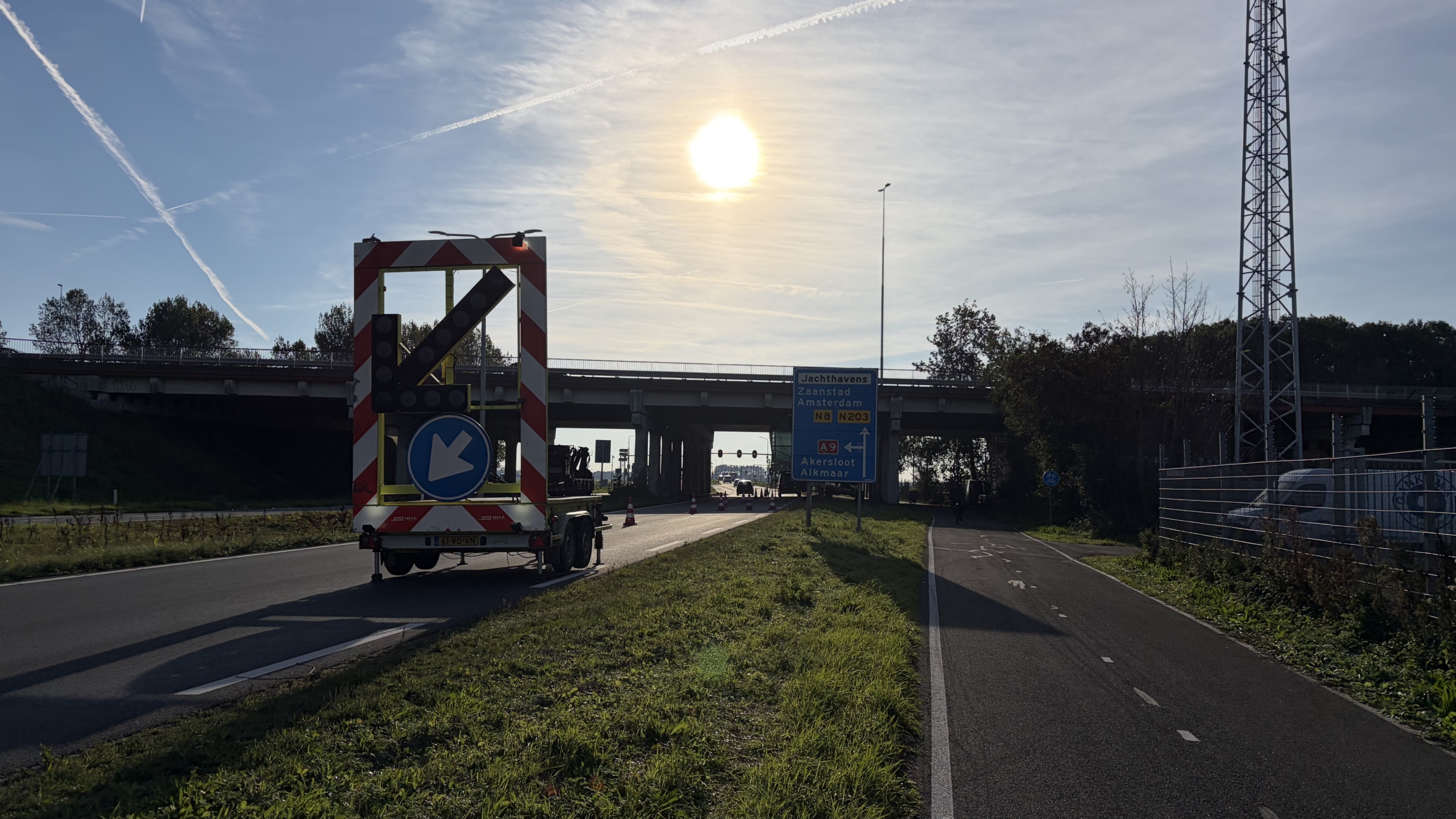 Pijlwagen met linksafpijl bij een viaduct en wegwijzer naar Jachthavens, Zaanstad en Amsterdam, met laagstaande zon