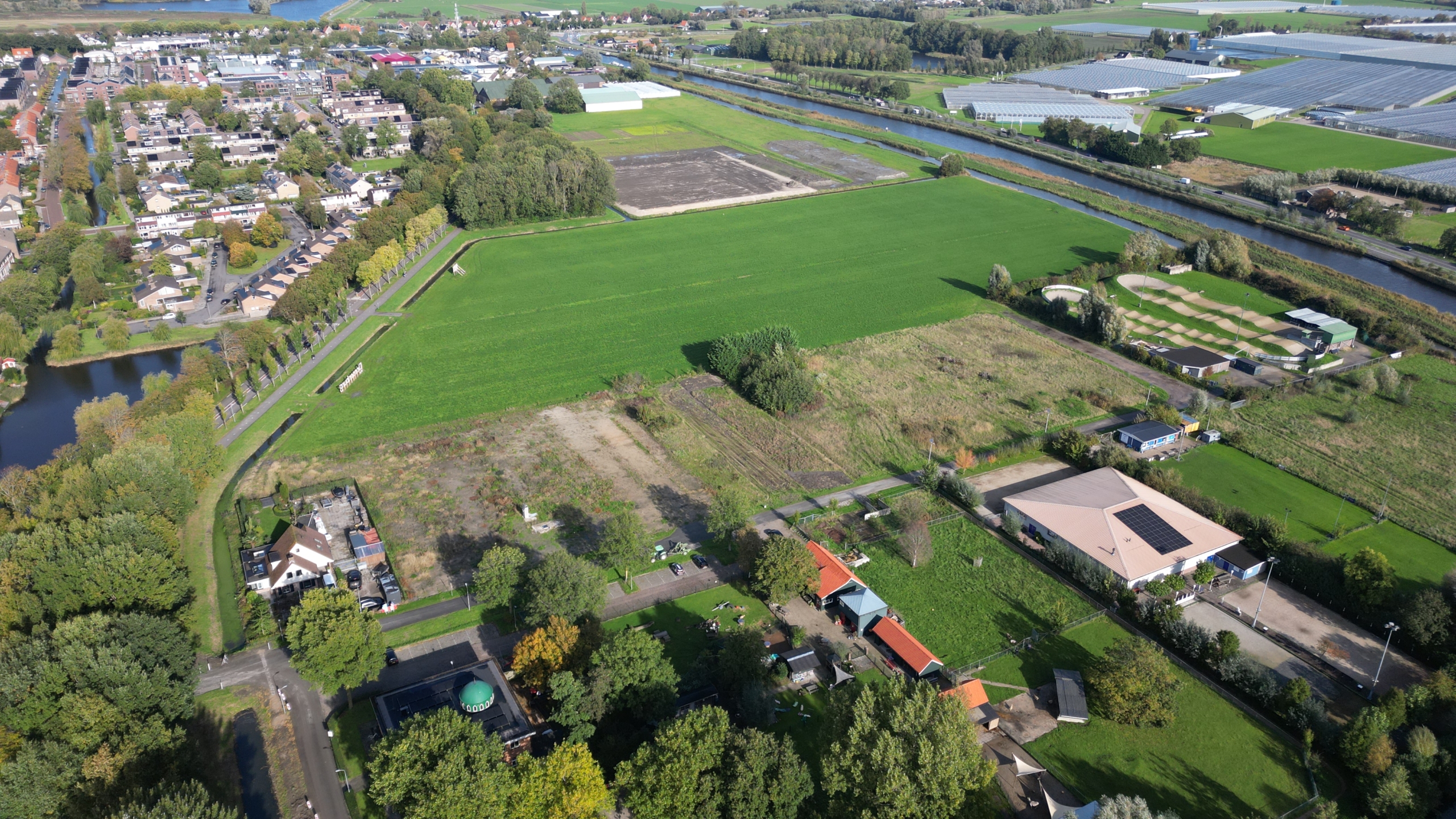 Luchtfoto van woonwijk en groene weilanden langs een kanaal, met kassen en sportvelden in de verte