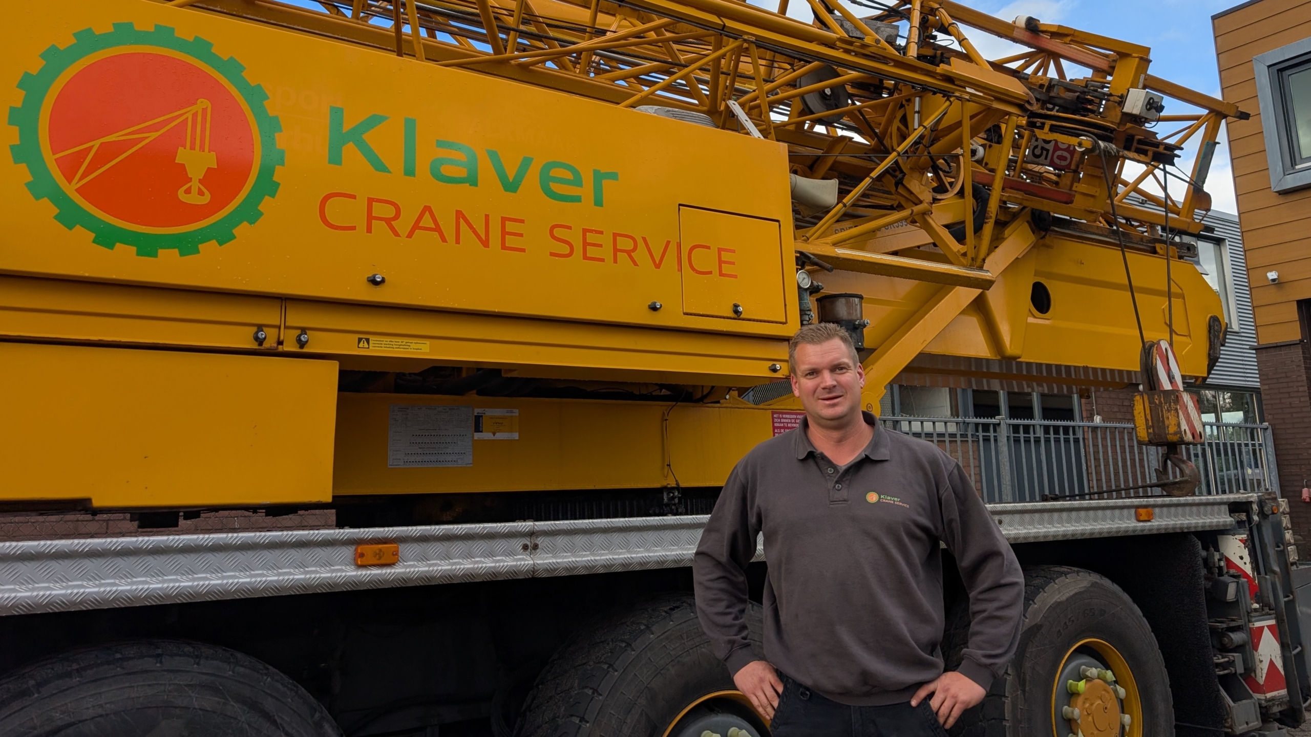 Man staat voor een gele mobiele kraan met het opschrift Klaver Crane Service, bij een bedrijfspand