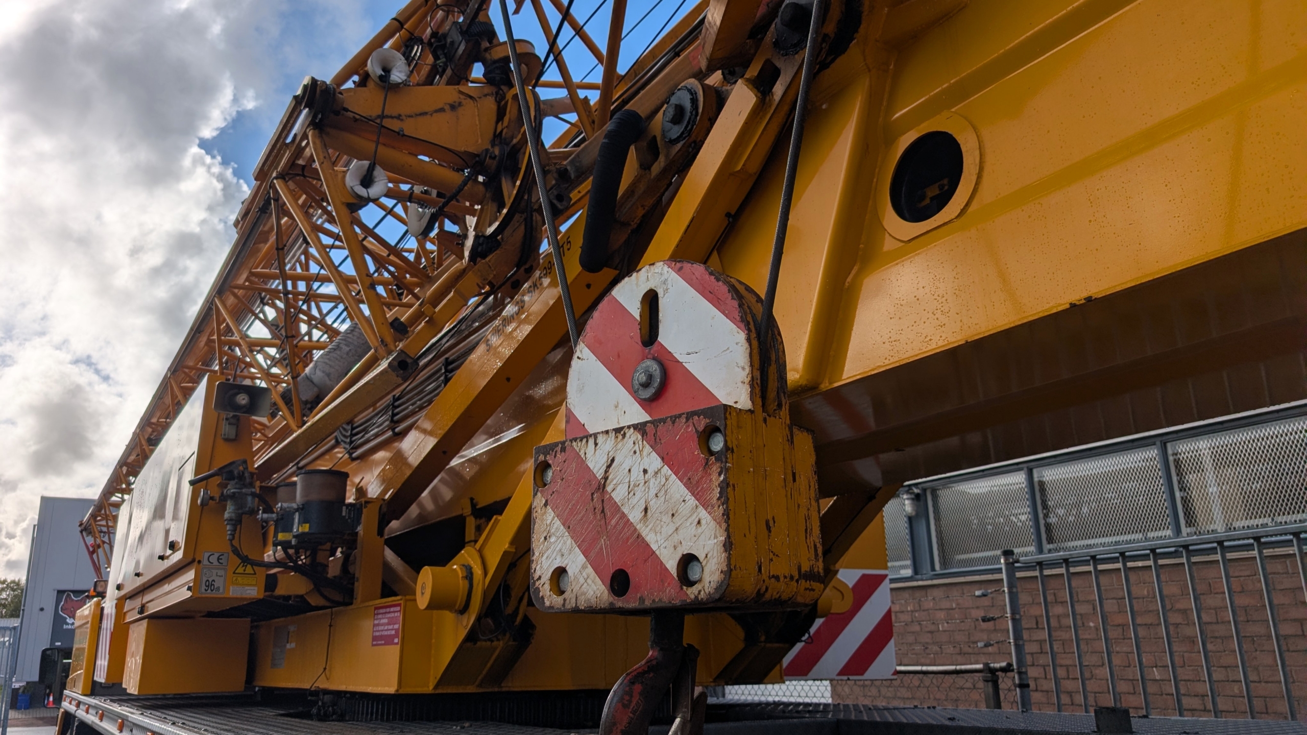 Detail van een gele hijskraan met hijshaak en rood-wit waarschuwingsbord, tegen een bewolkte lucht