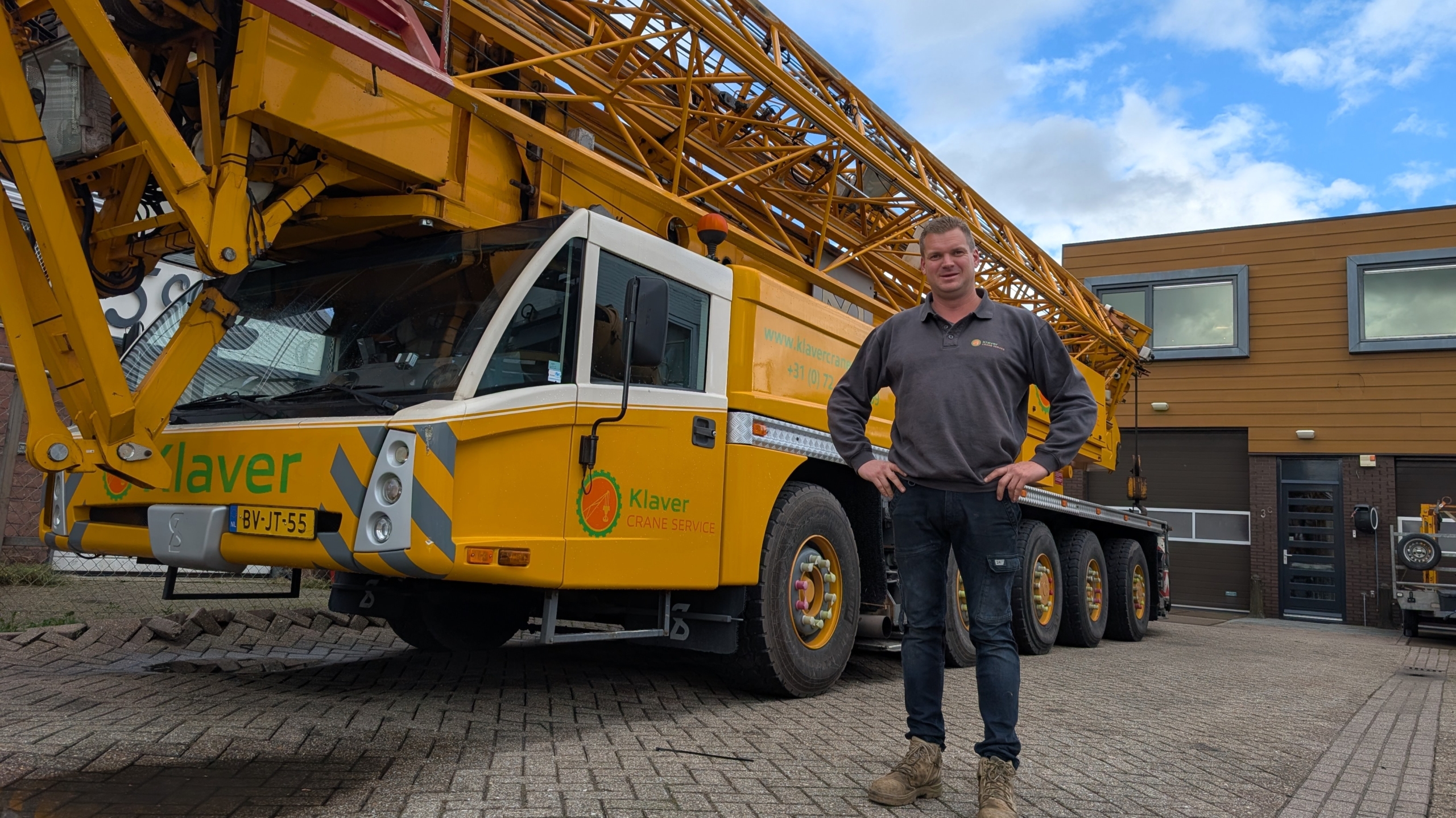 Man staat met handen in de zij naast een grote gele mobiele hijskraan van Klaver Crane Service bij een bedrijfspand