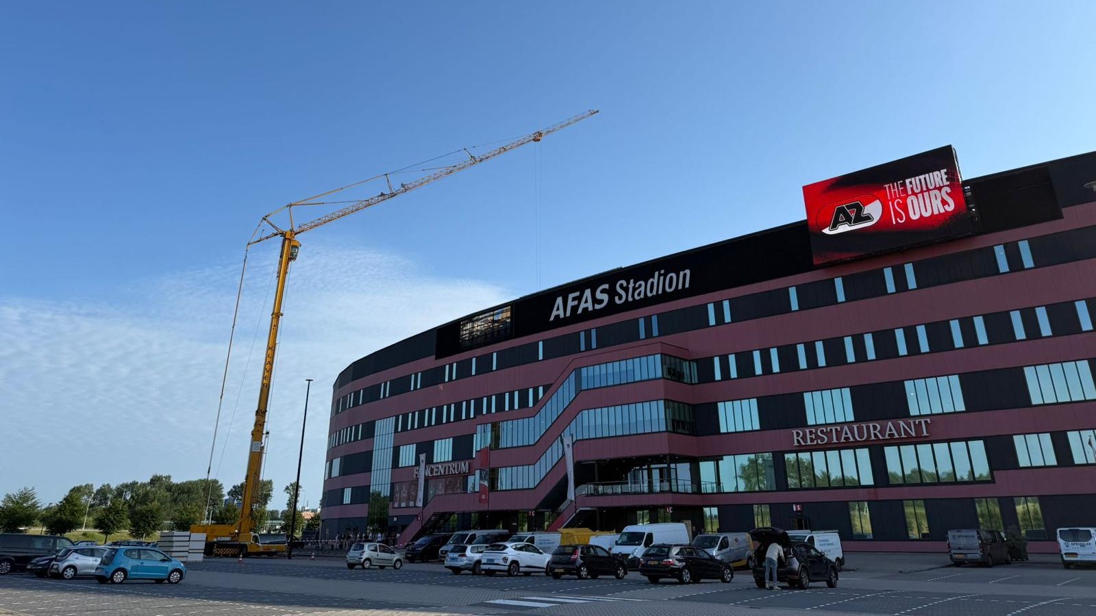 Buitenkant van AFAS Stadion met grote bouwkraan, parkeerterrein met auto’s en bord met tekst “THE FUTURE IS OURS”
