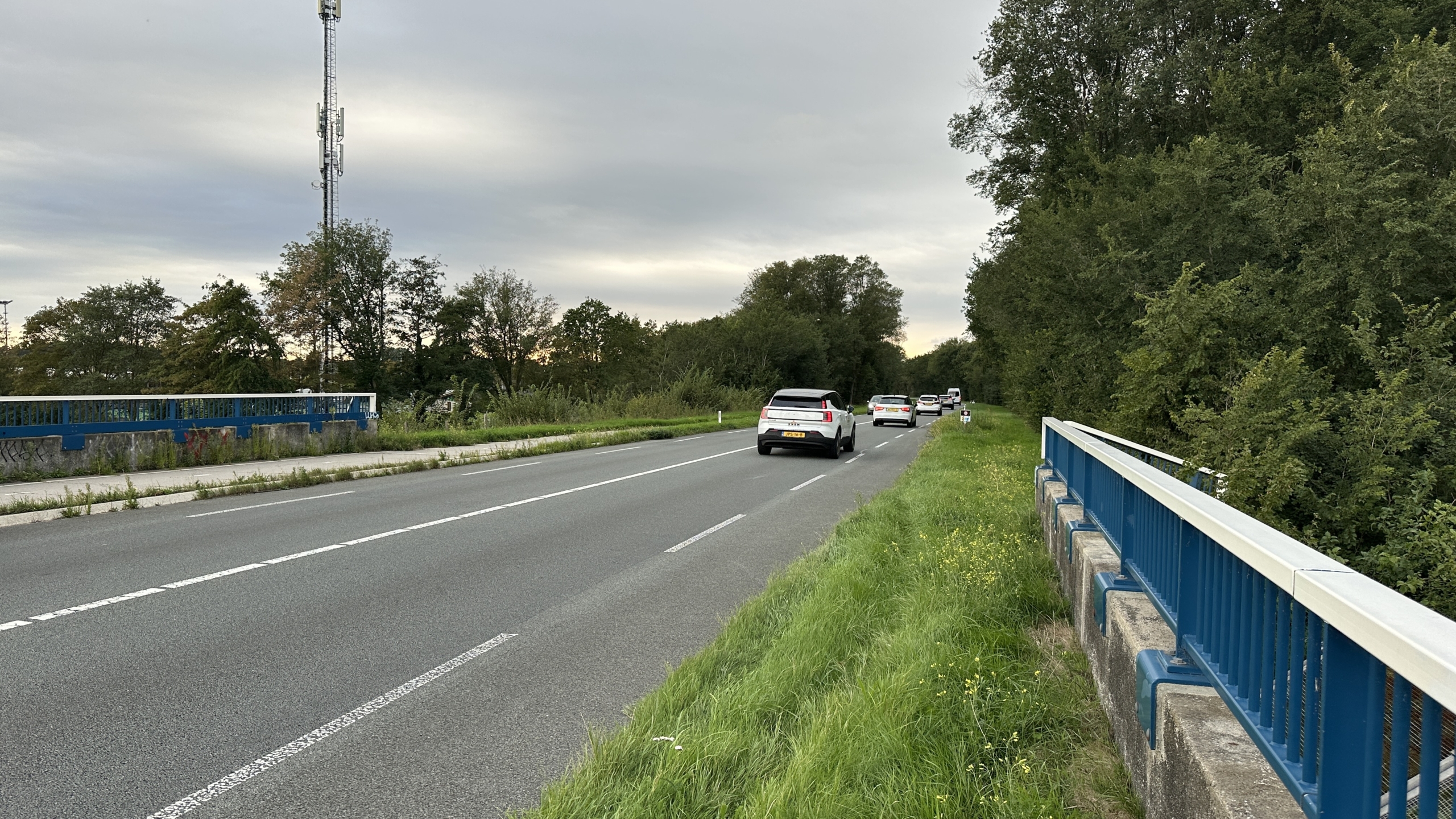 Landweg met auto’s op rij langs groen en bomen, met blauwe brugleuningen en een zendmast onder bewolkte lucht