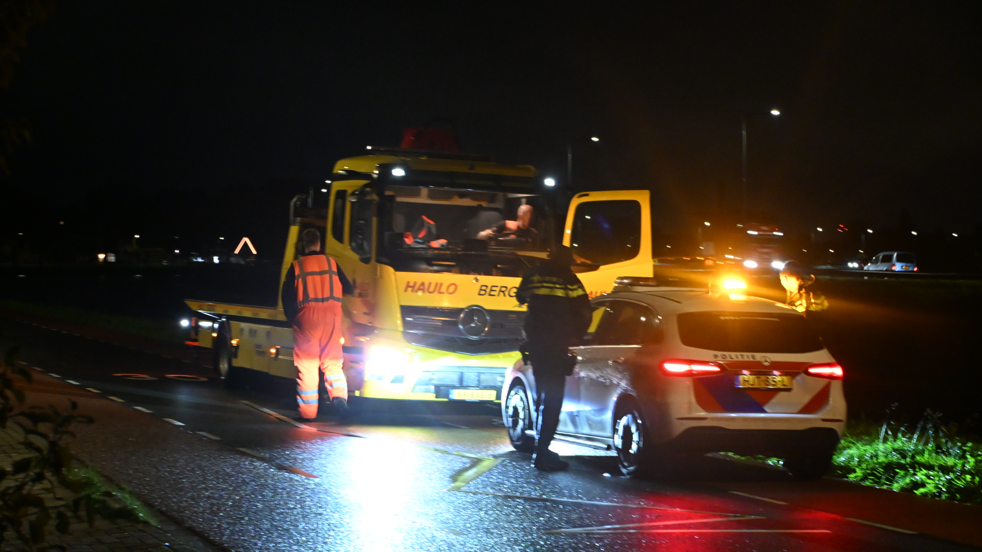Bergingswagen en politieauto met zwaailichten op natte weg in de nacht, met hulpverleners ernaast