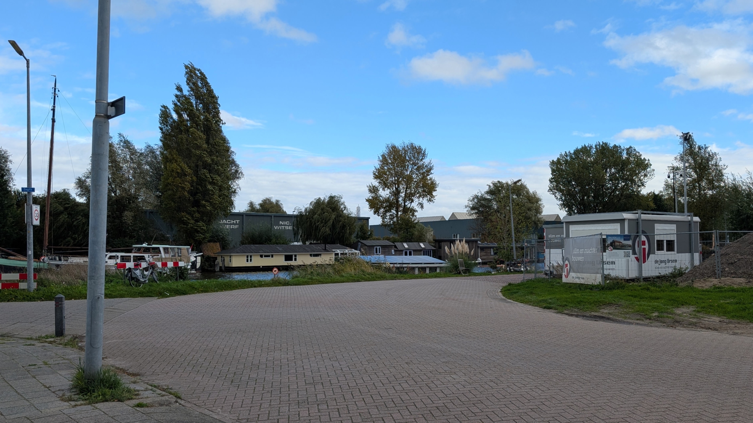 Lege weg langs een kanaal met woonboten, bomen en een bouwplaats met keet en hekwerk