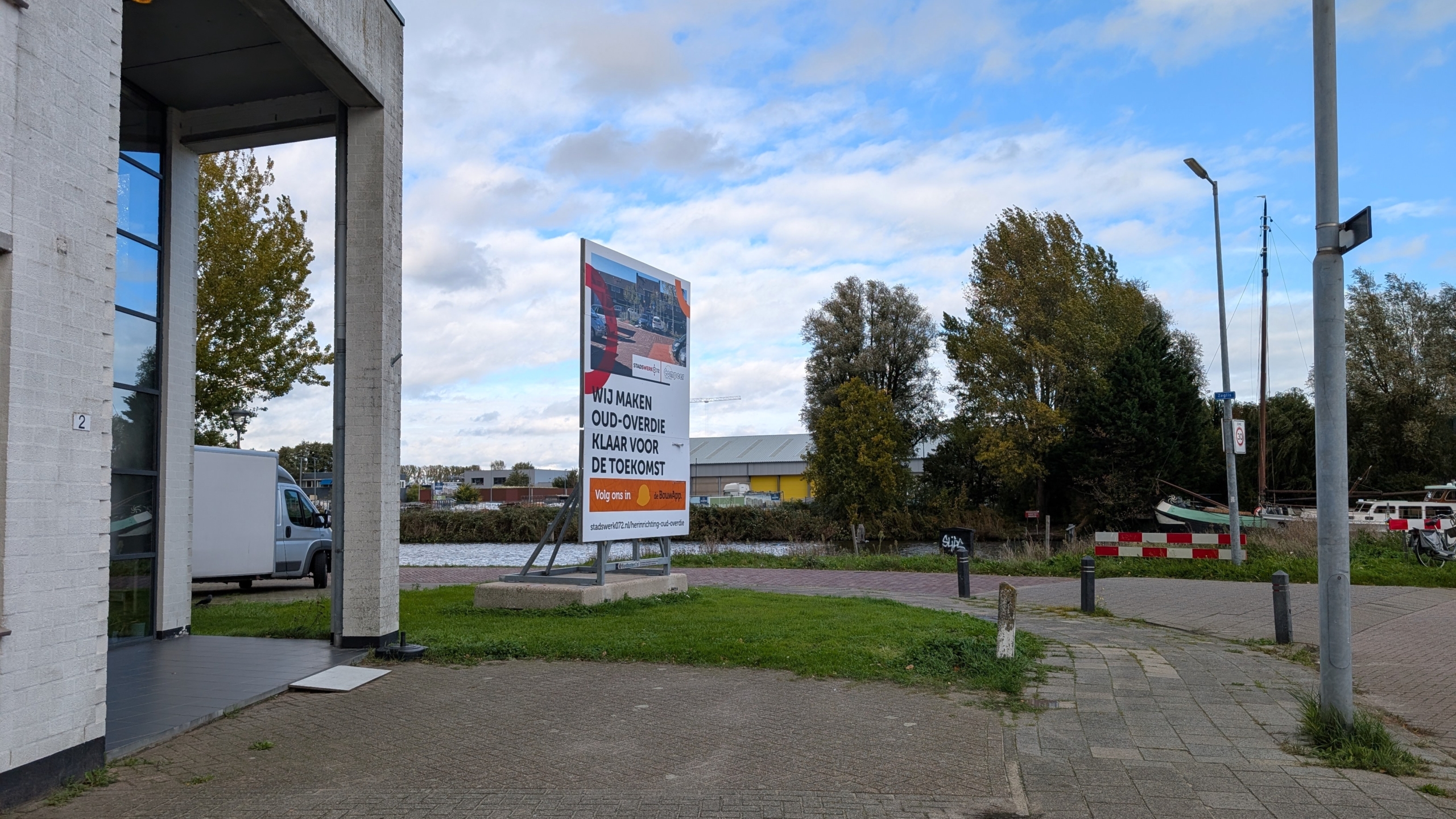 Informatiebord langs het water met tekst “Wij maken Oud-Overdie klaar voor de toekomst” naast een gebouw en weg