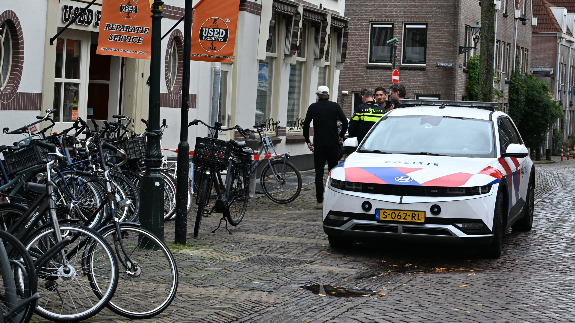Politieauto in smalle straat met fietsenrekken; agenten en omstanders praten bij rood-wit afzetlint voor een winkel