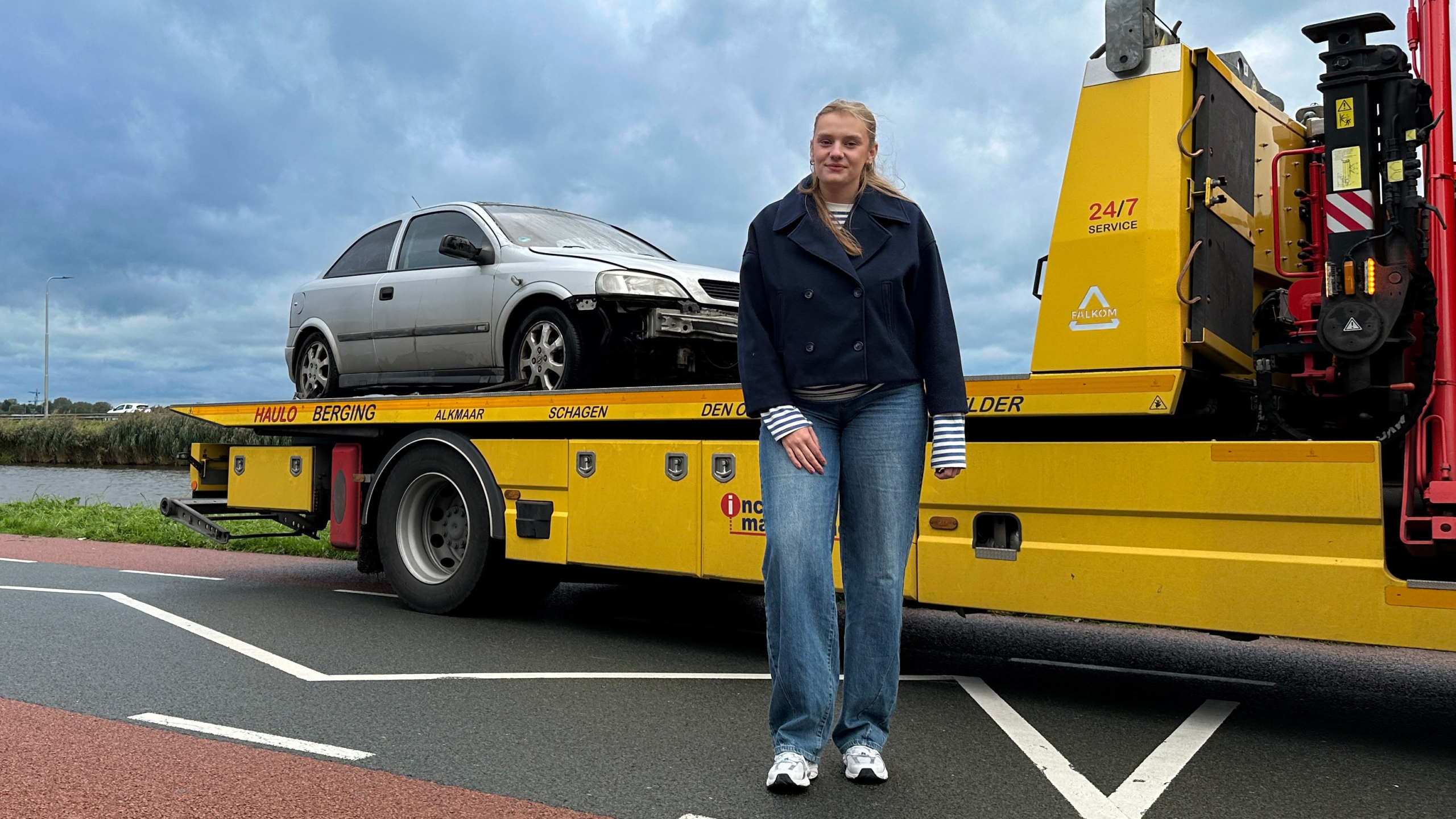 Vrouw staat op de weg naast een gele bergingswagen met een beschadigde zilveren auto op het platform