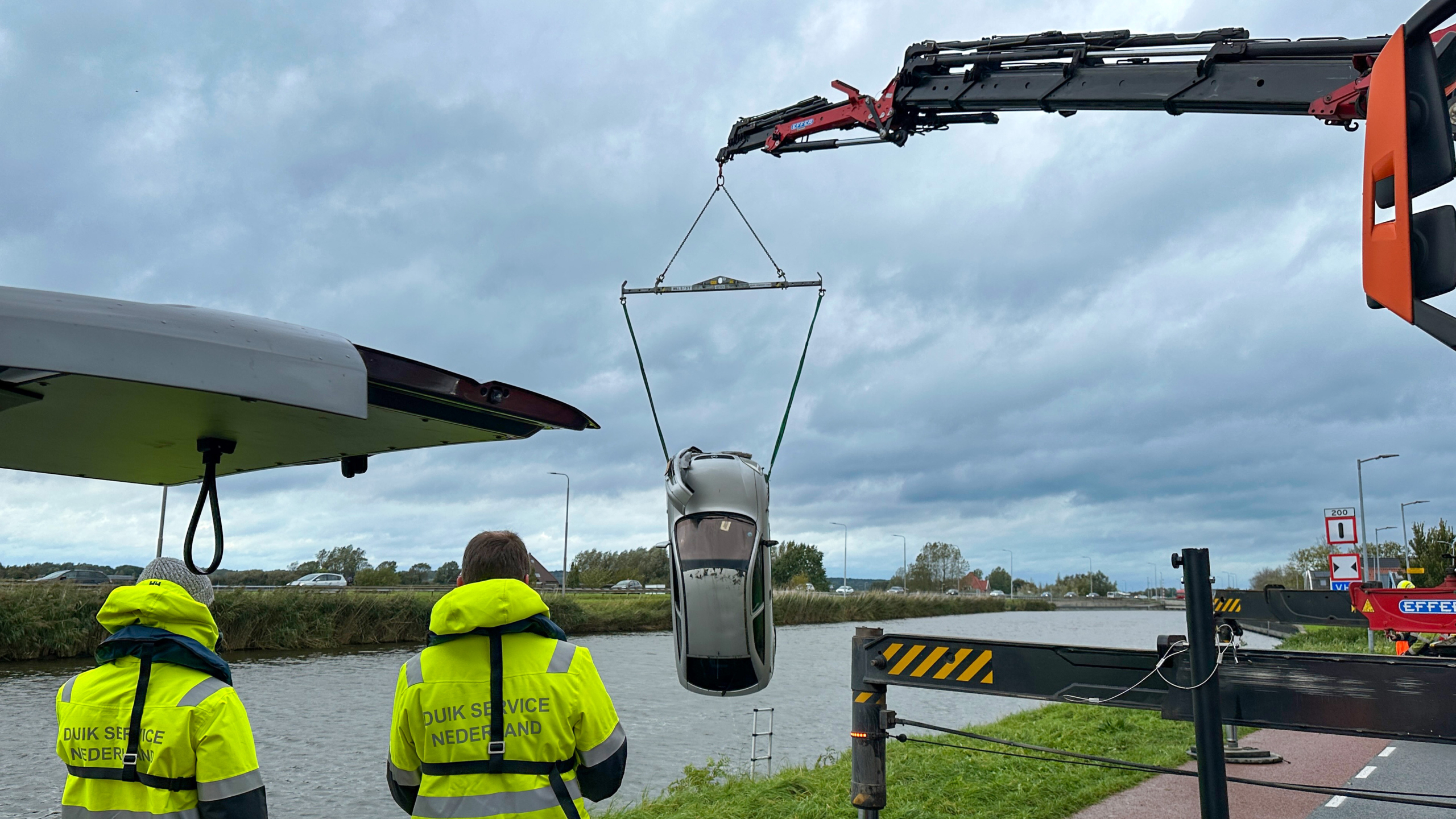 Kraan hijst auto uit een kanaal terwijl twee duikers in gele jassen toekijken onder een bewolkte lucht