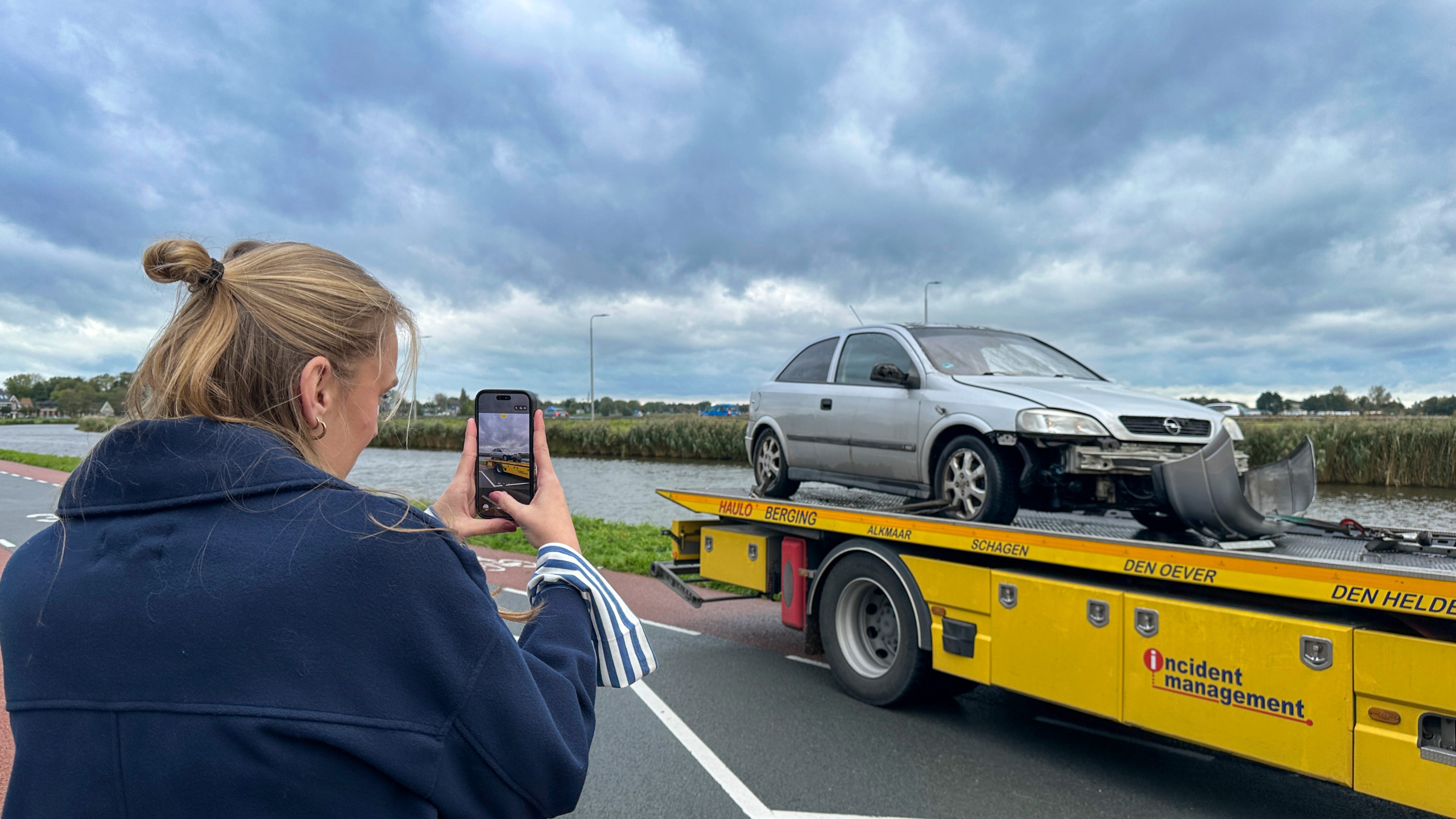 Vrouw fotografeert met haar telefoon een beschadigde auto op een gele bergingswagen langs het water onder bewolkte lucht