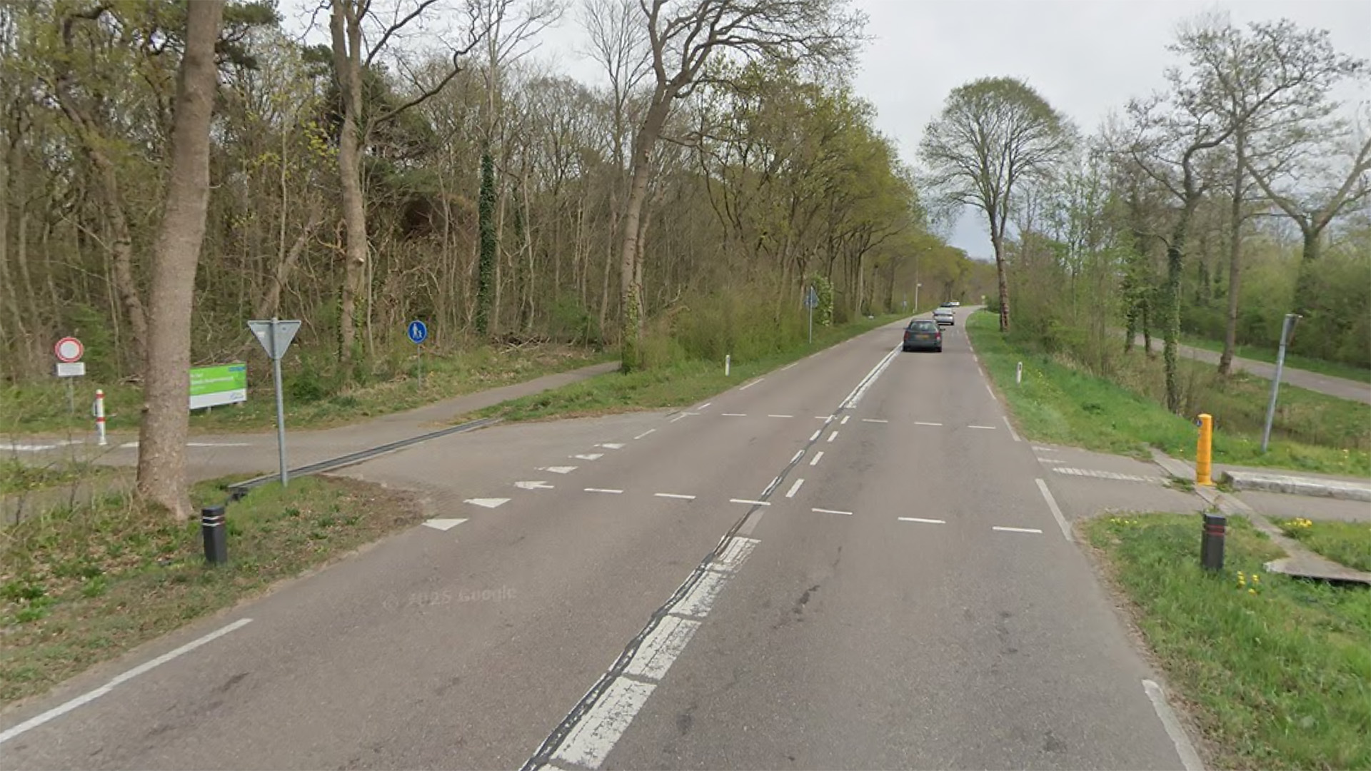 Landweg door bosrijk gebied met zijweg en fietspad, enkele auto’s in de verte en verkeersborden langs de berm