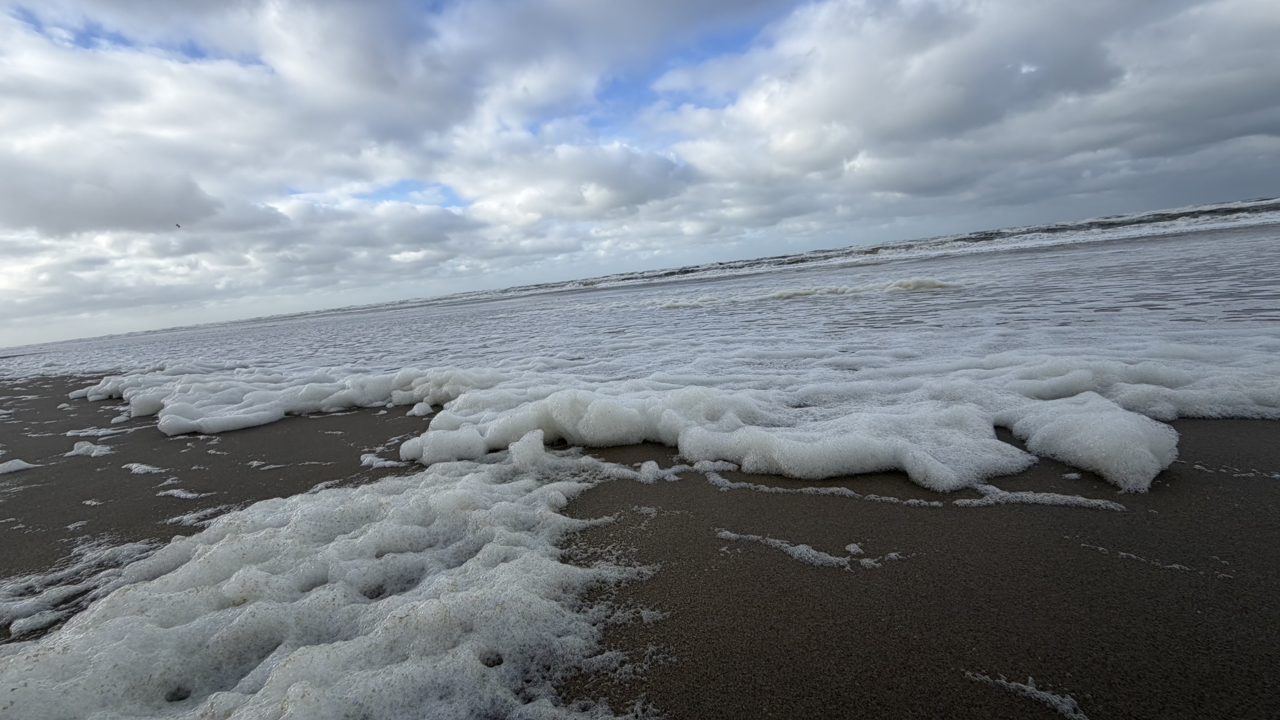 Schuim van de branding op het strand, met ruige zee en bewolkte lucht met stukjes blauw