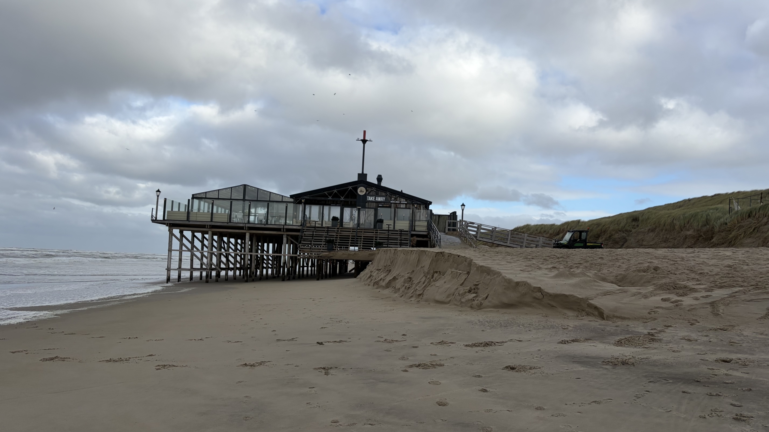 Strandpaviljoen op palen aan zee met glazen serre en bordje Take Away, onder bewolkte lucht en duinen