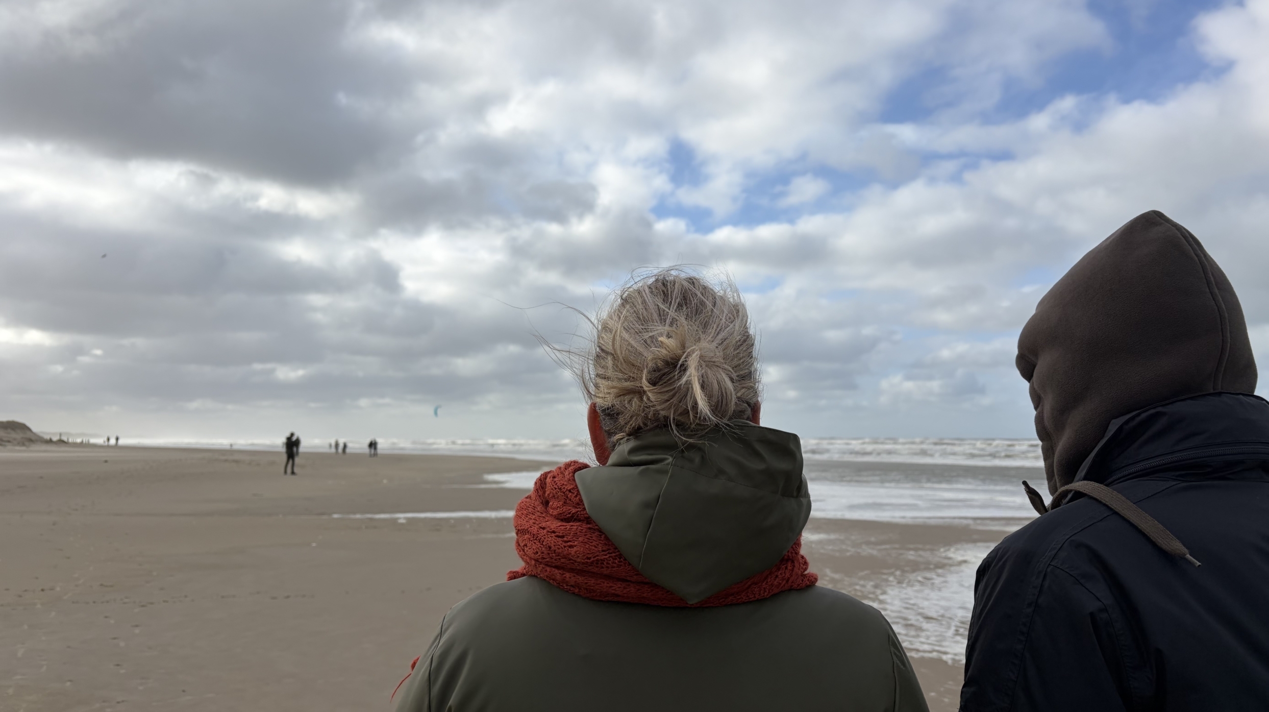 Twee personen in winterjassen, van achteren gezien, kijken uit over een winderig strand met wolkenlucht en zee