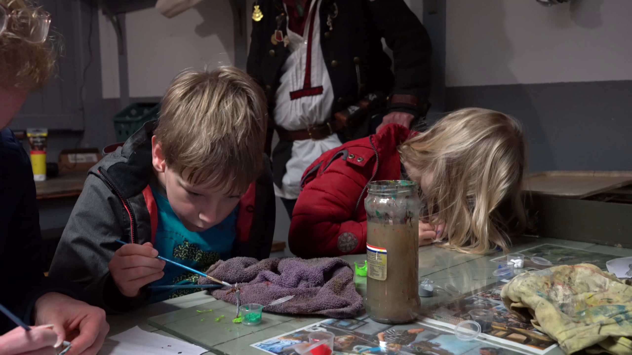 Twee kinderen schilderen geconcentreerd aan een tafel met pot verfwater, kwasten en verfspatten, terwijl een volwassene toekijkt