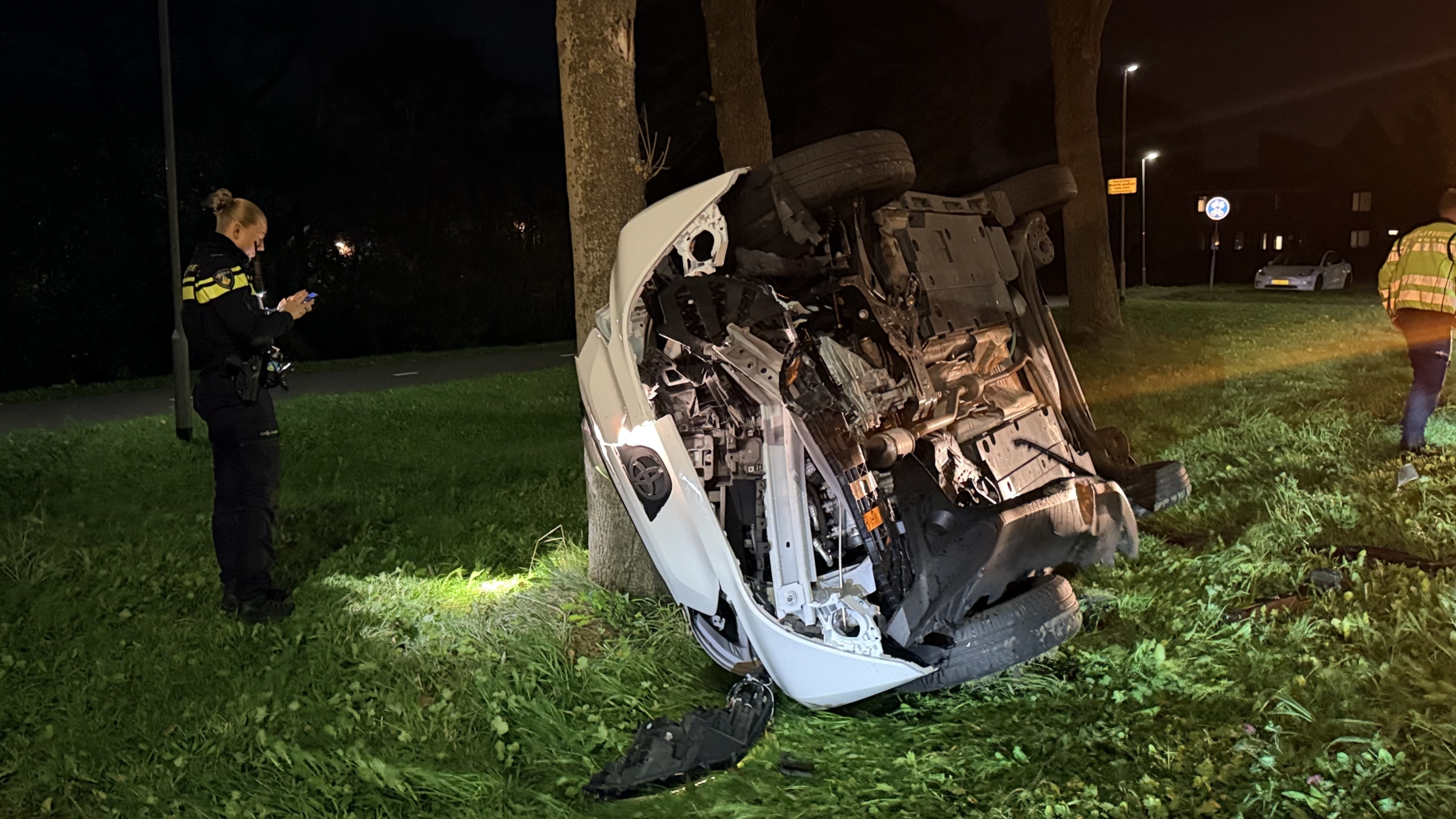 Omgeslagen auto tegen een boom op een grasveld bij nacht, met politieagent en hulpverlener in de buurt