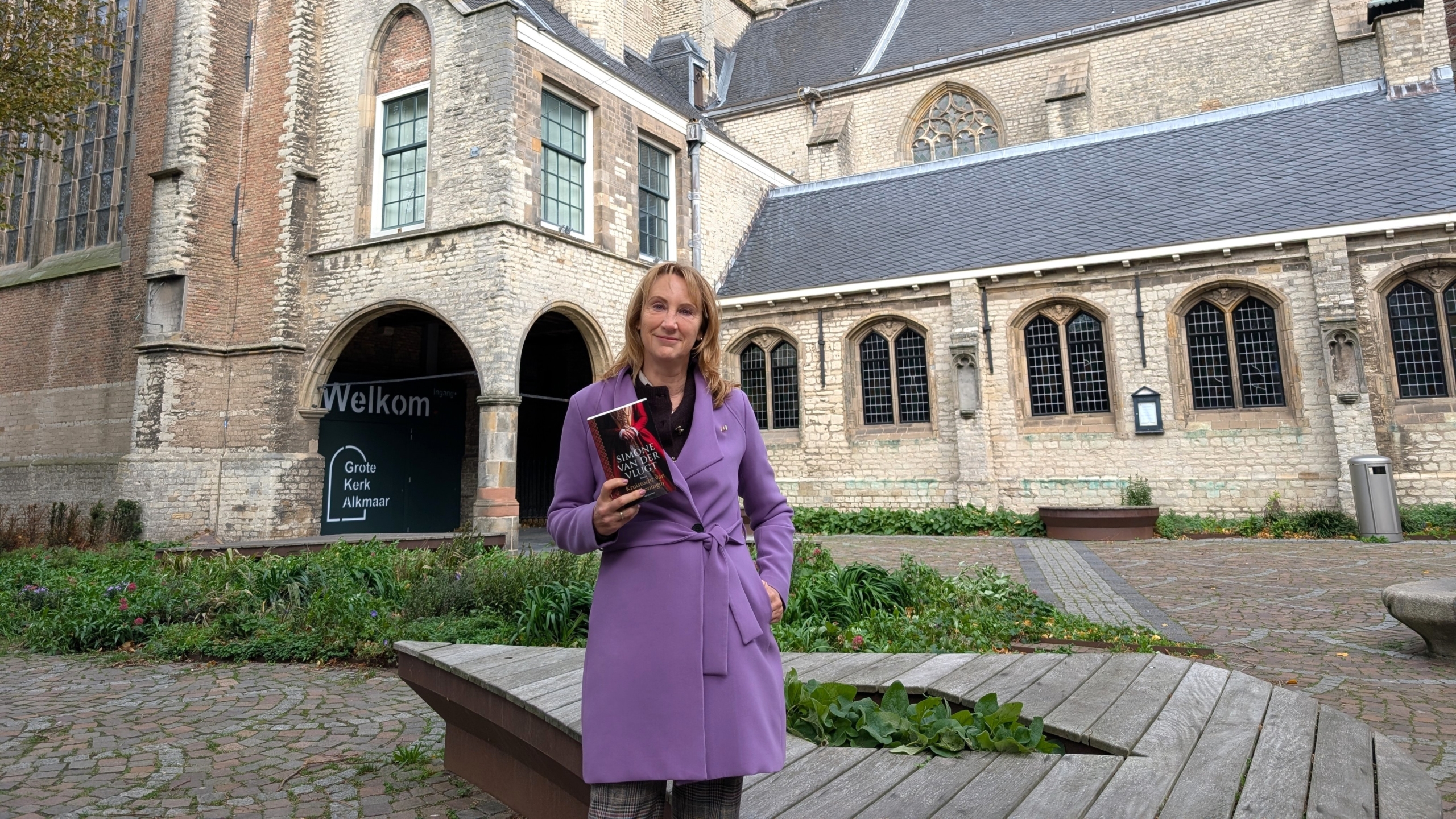 Vrouw in paarse jas houdt een boek vast voor de ingang van de Grote Kerk Alkmaar met bordje “Welkom”