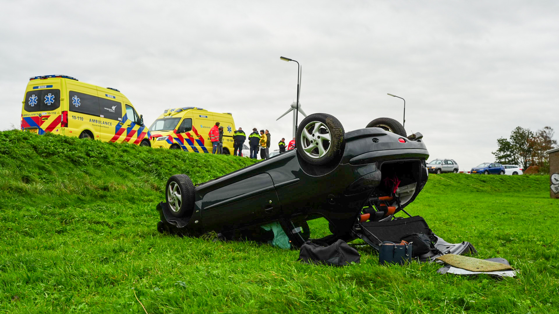 Omgeslagen auto op een grashelling, met ambulances en hulpdiensten op de weg erboven