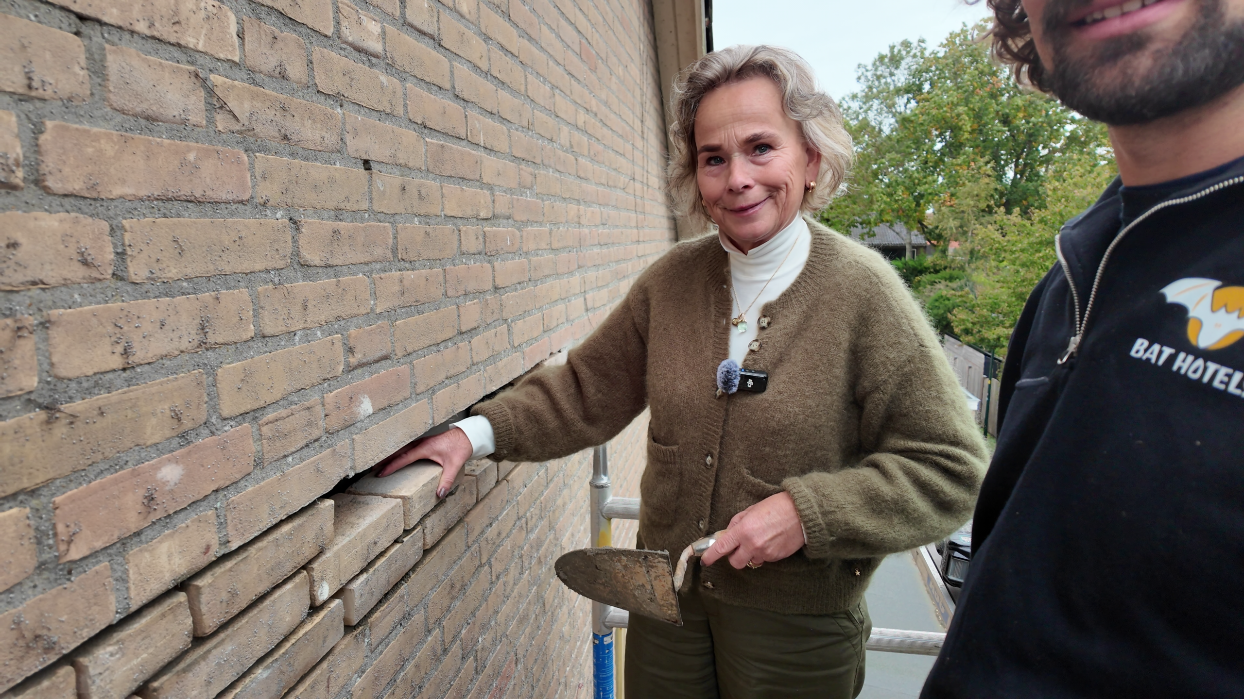 Vrouw op steiger bij bakstenen gevel, controleert loszittend metselwerk en houdt een metseltroffel vast