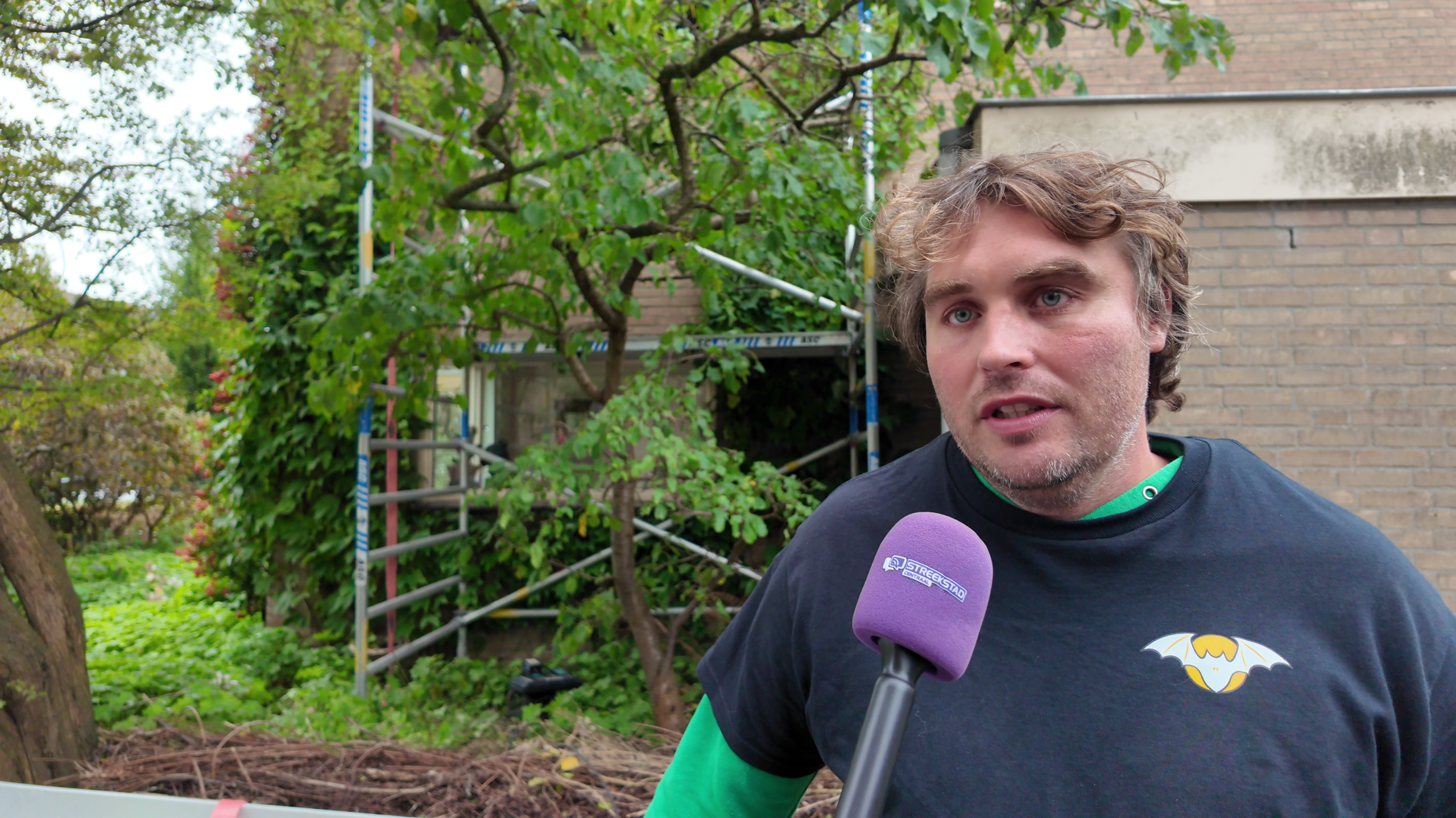Man wordt geïnterviewd met paarse microfoon voor een bakstenen gebouw met steiger en bomen op de achtergrond