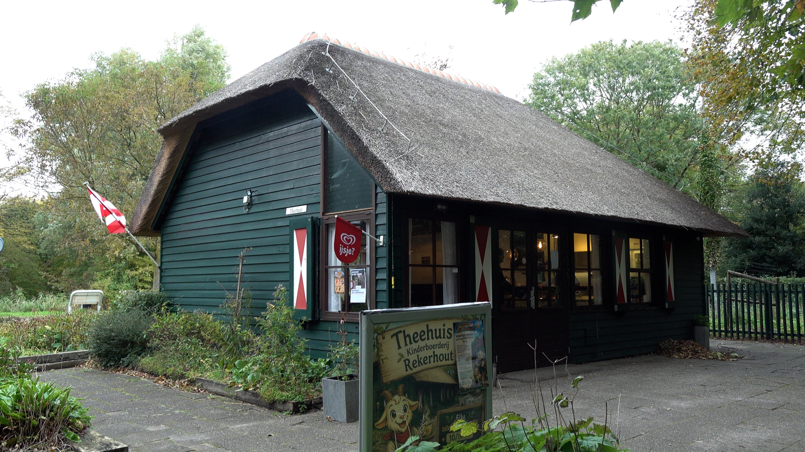 Groen houten theehuis met rieten dak en rood-witte luiken, met bord Theehuis Kinderboerderij Rekerhout