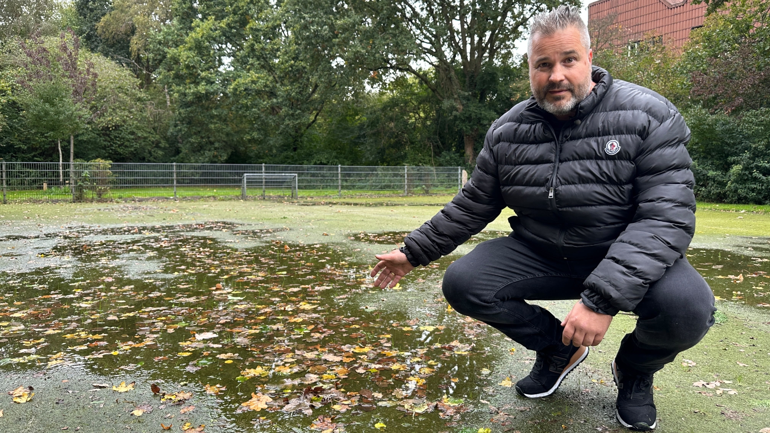 Man in zwarte gewatteerde jas hurkt bij een plas met bladeren op een groen veld, met bomen en hek op de achtergrond