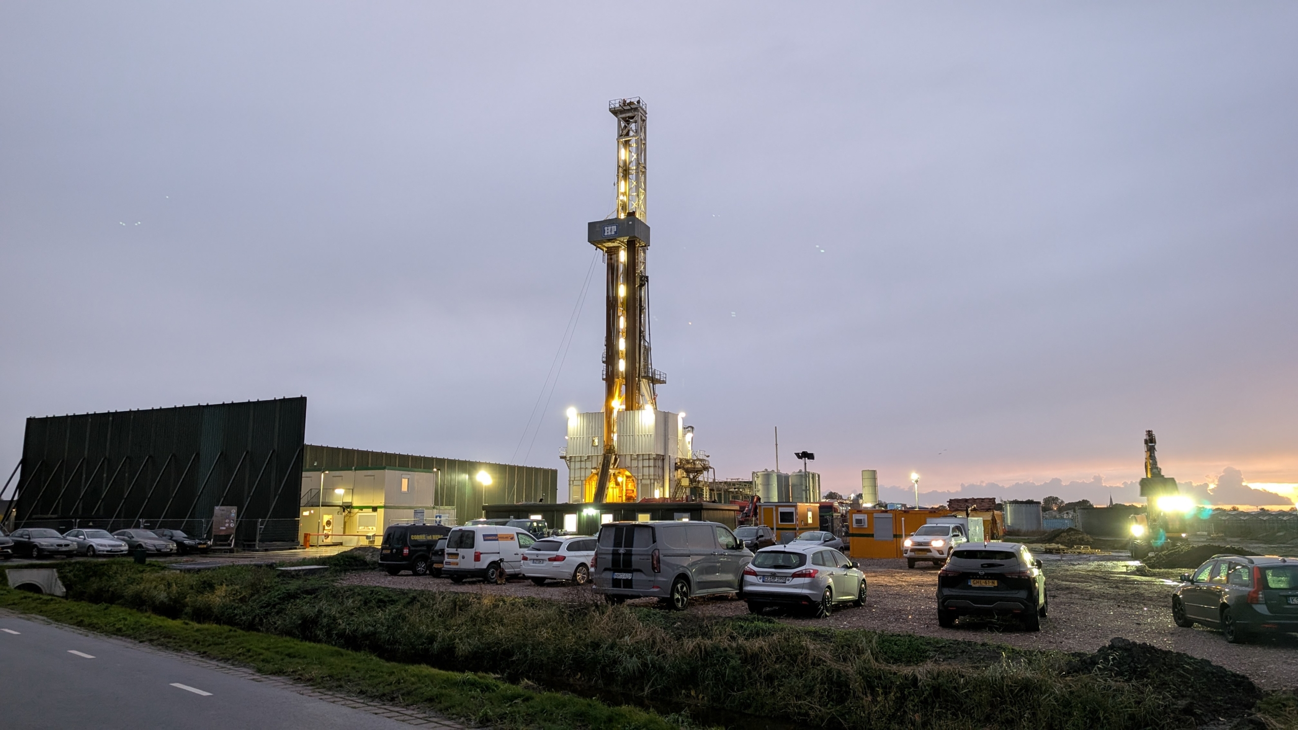 Verlichte boortoren op een industrieterrein bij schemering, met geparkeerde auto’s en bouwcontainers op de voorgrond
