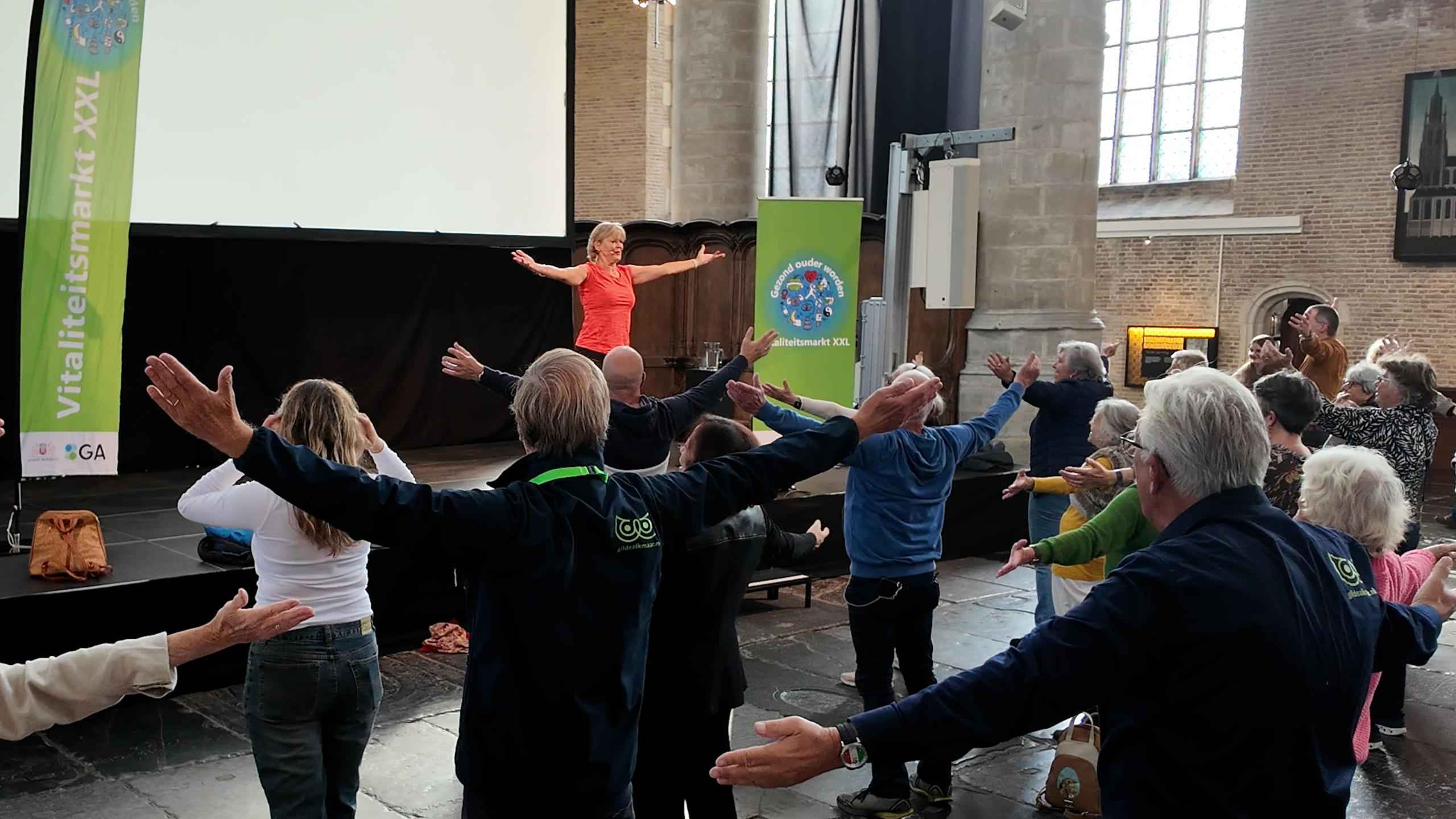 Groep mensen doet gezamenlijke oefeningen met armen wijd, begeleid door instructeur op podium in grote hal