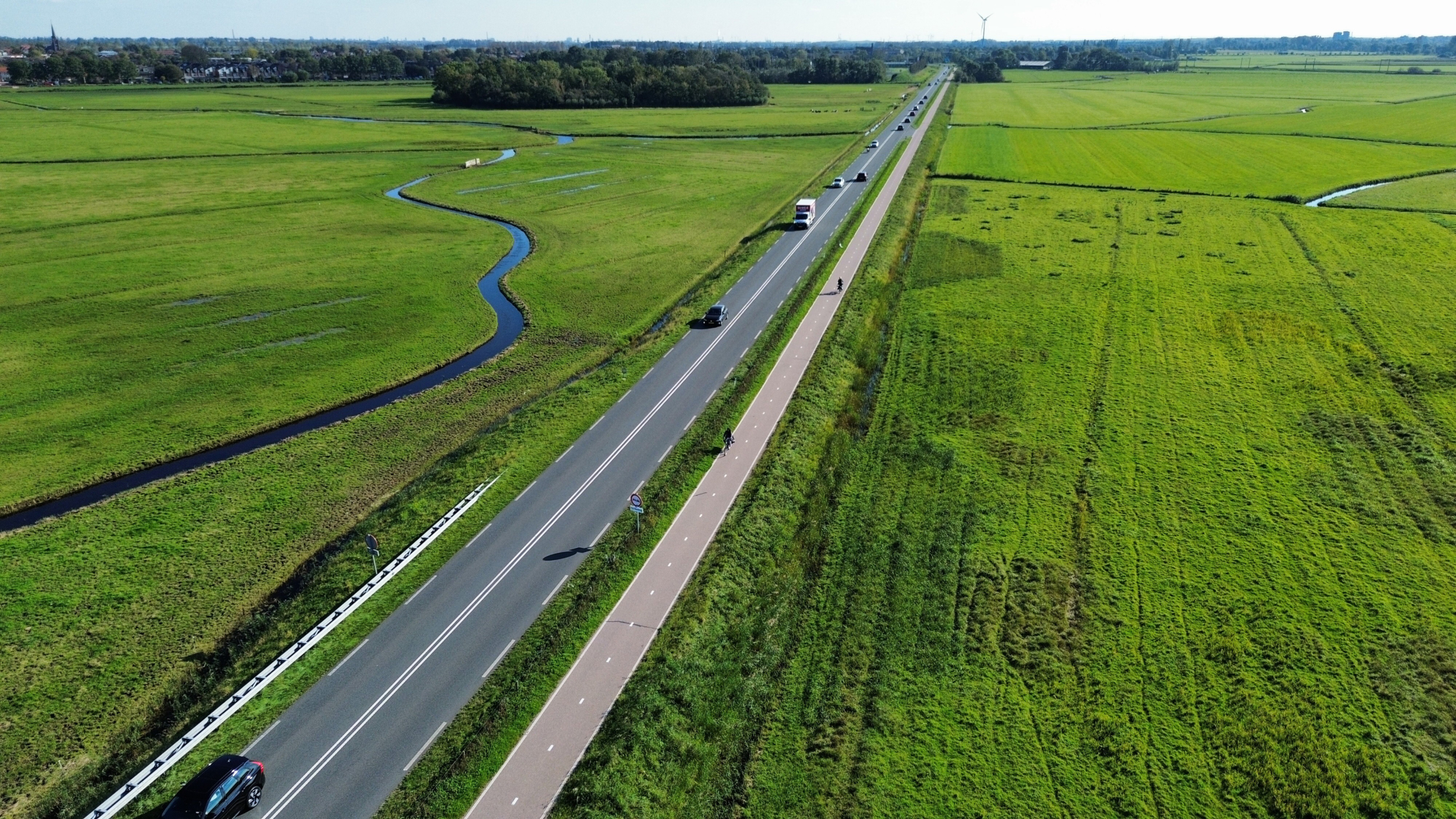 Luchtfoto van een lange weg met fietspad langs groene weilanden en een slingerende sloot, met auto’s en fietsers