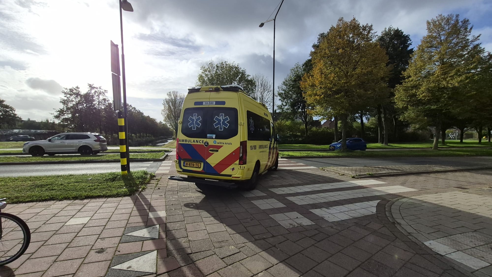 Gele ambulance van Witte Kruis geparkeerd bij een zebrapad langs een weg met bomen en auto’s