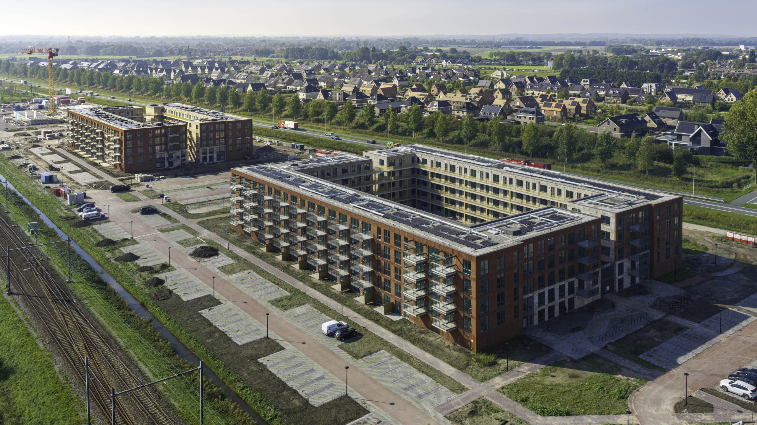Luchtfoto van nieuwbouwappartementen met balkons naast spoorlijn, met woonwijk en groen op de achtergrond