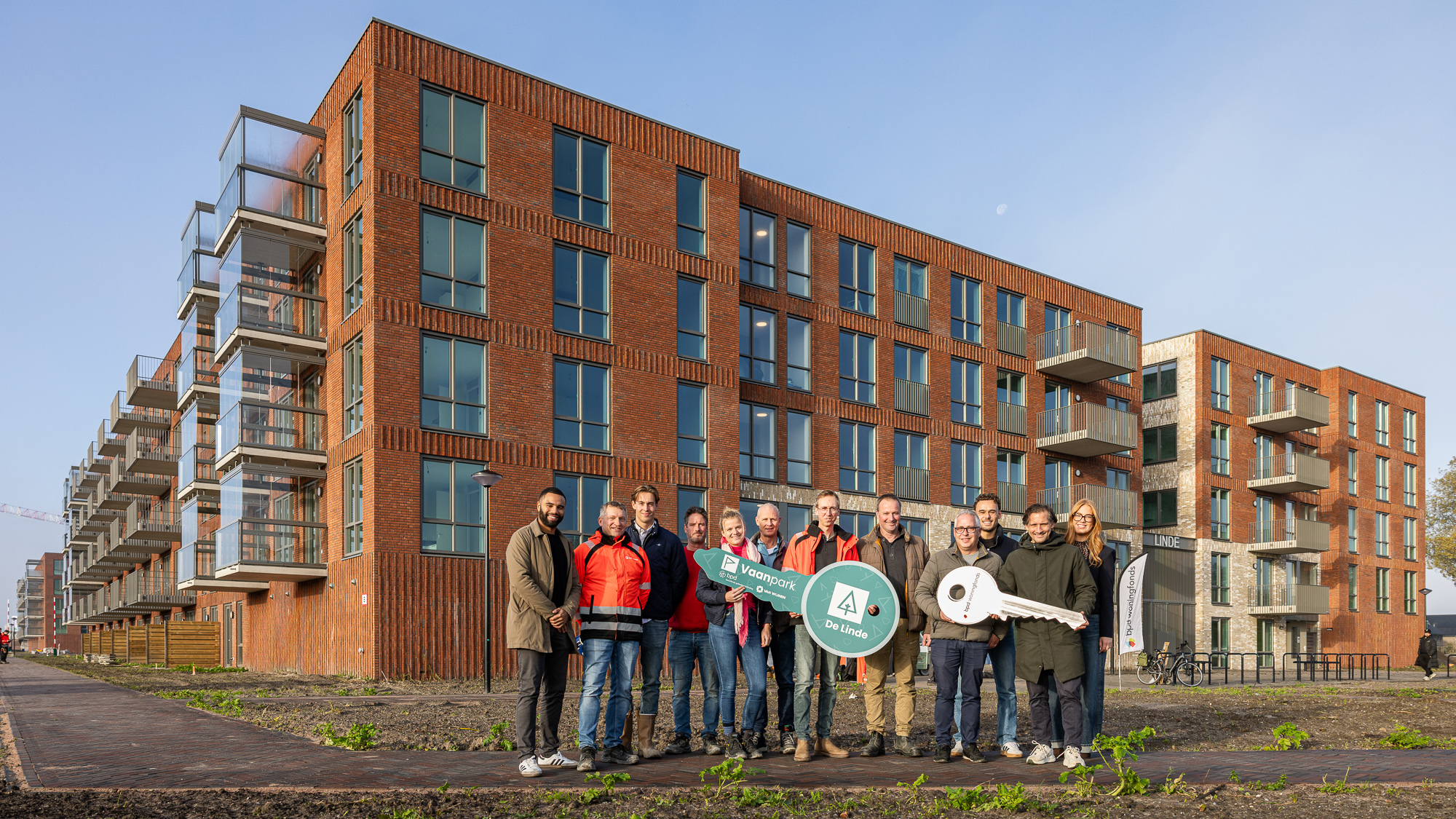 Groep mensen voor nieuw bakstenen appartementencomplex, met borden “Vaanpark De Linde” en een grote symbolische sleutel