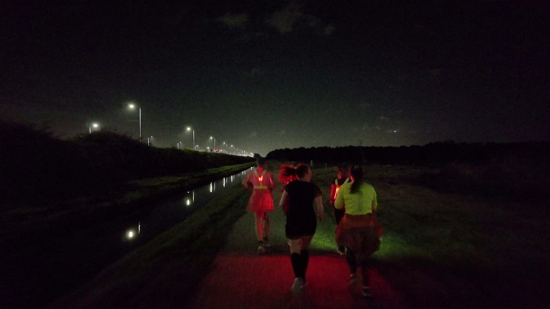 Groep hardlopers met rode lampjes op een donker pad langs een kanaal, met straatverlichting in de verte