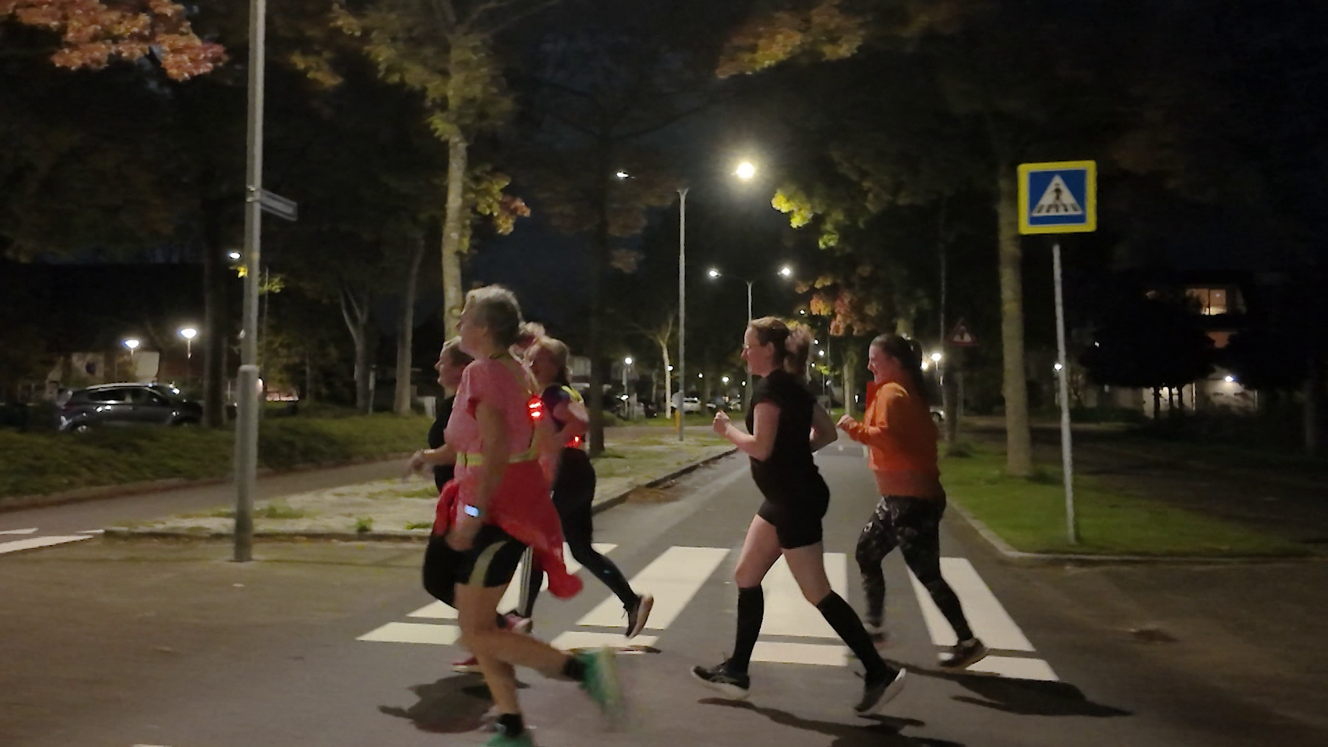 Groep hardlopers steekt ’s avonds een zebrapad over op een verlichte, met bomen omzoomde straat