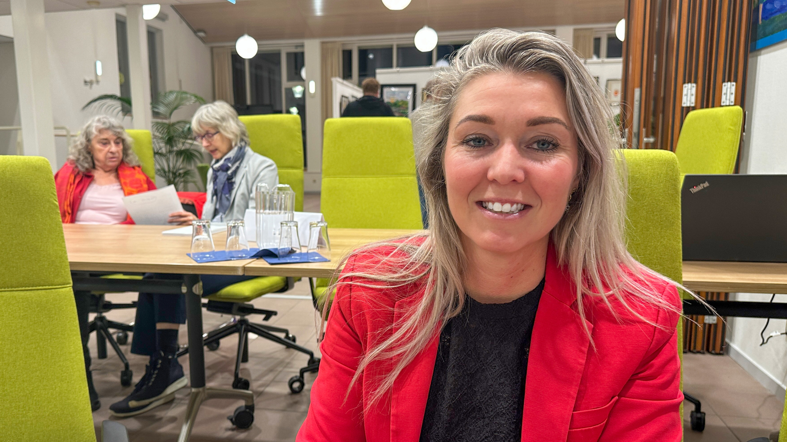 Lachende vrouw in rood jasje op de voorgrond in vergaderruimte met groene stoelen en twee lezende mensen op de achtergrond