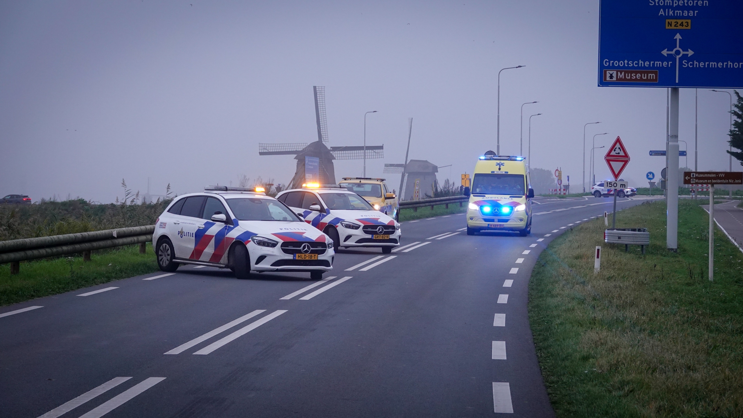 Twee politieauto’s en een ambulance met zwaailichten op een landelijke weg, met molens op de achtergrond