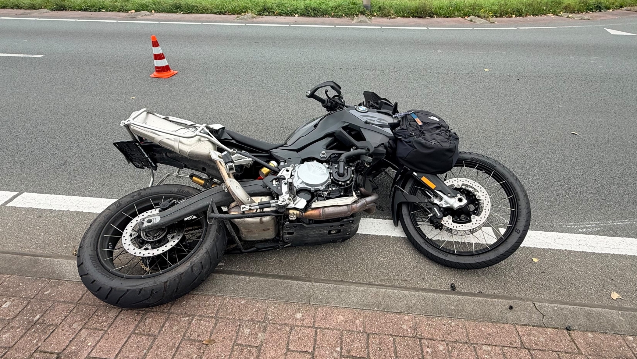 Omgevallen motorfiets op de rijbaan naast een fietsstrook, met verkeerskegel en auto’s op de achtergrond