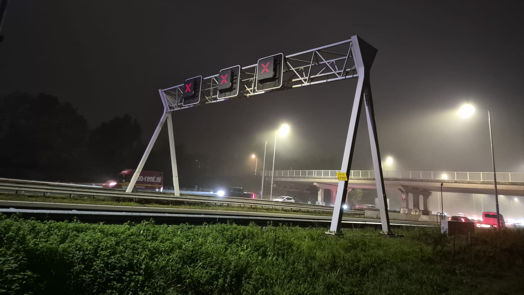 Portaal boven de snelweg met drie rijstrooksignalen met rode kruisen, bij nachtelijke mist en straatverlichting