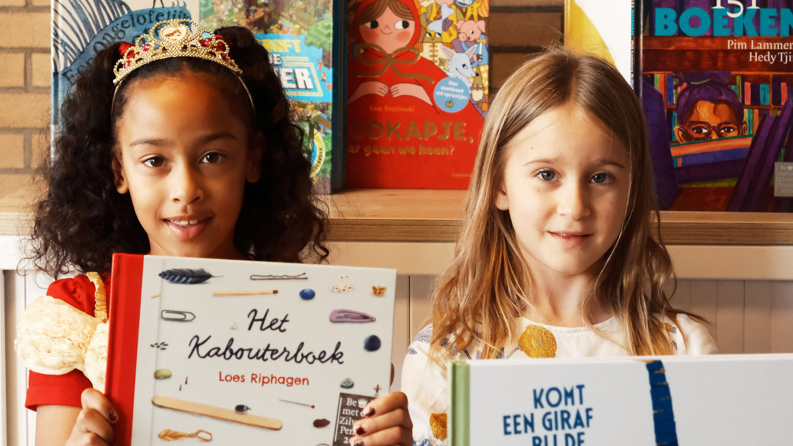 Twee meisjes houden kinderboeken omhoog voor een boekenkast; links draagt een meisje een kroon, rechts een blond meisje