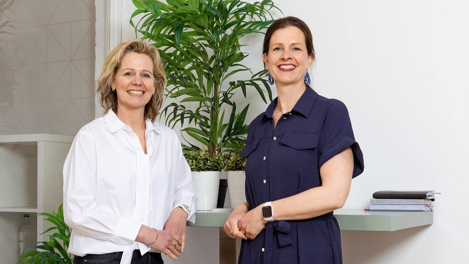 Twee glimlachende vrouwen staan in een kantoorruimte bij een balie, met grote groene kamerplant op de achtergrond