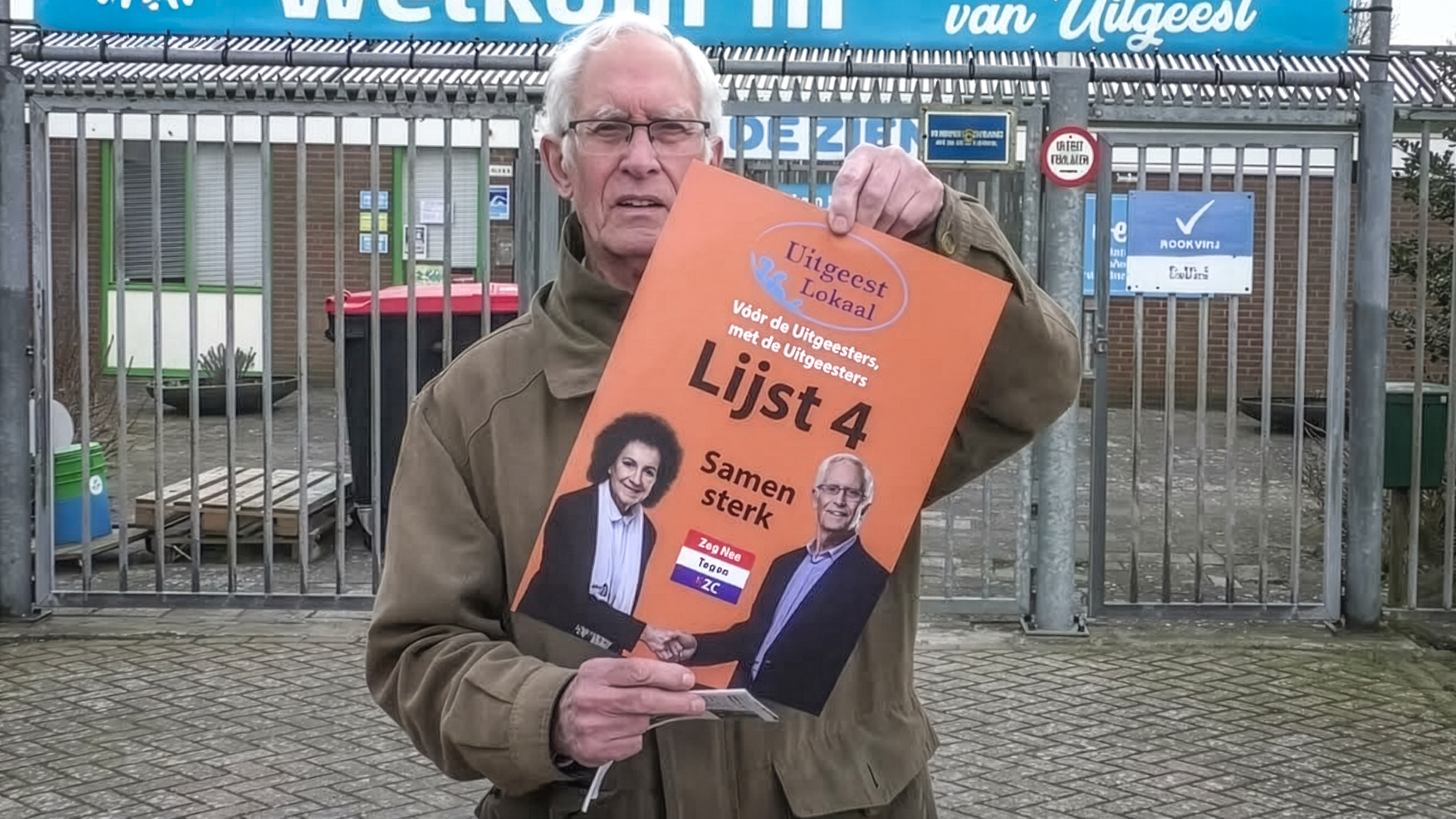 Oudere man houdt campagneposter vast met tekst Uilgeest Lokaal, Lijst 4 en Samen sterk, voor een hek