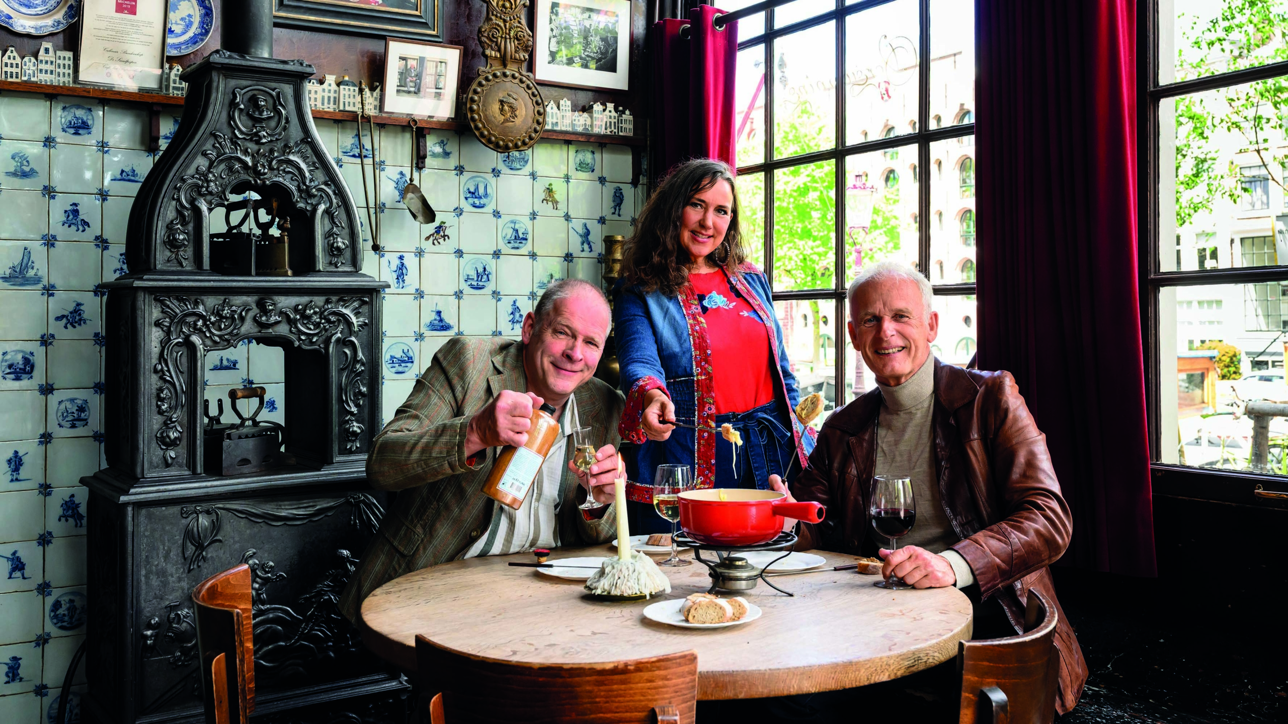 Drie mensen aan tafel in een gezellig restaurant met fonduepan, wijn en oud-Hollandse tegels op de muur
