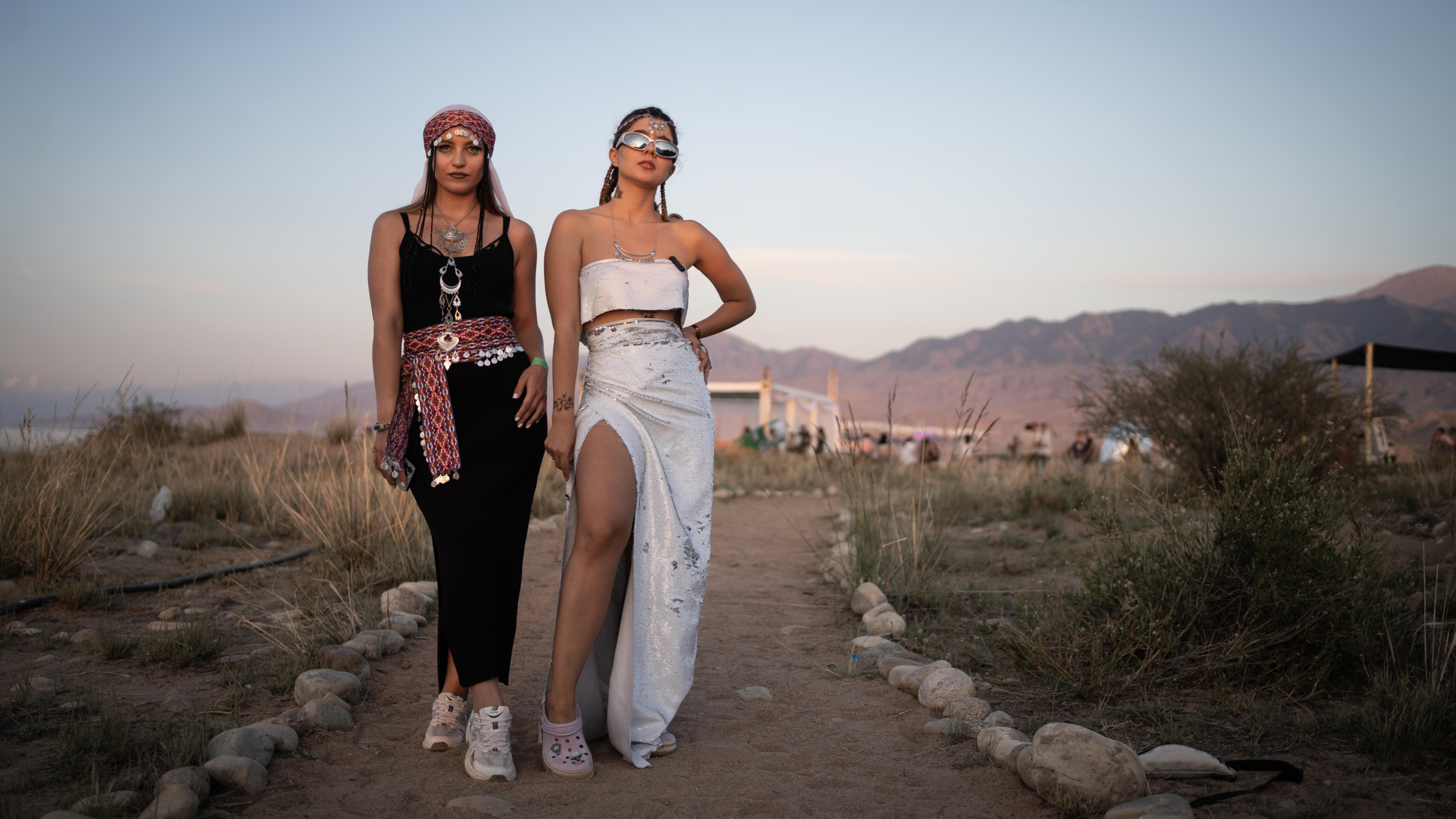 Twee vrouwen in opvallende festivaloutfits op een zandpad, met bergen en avondlucht op de achtergrond